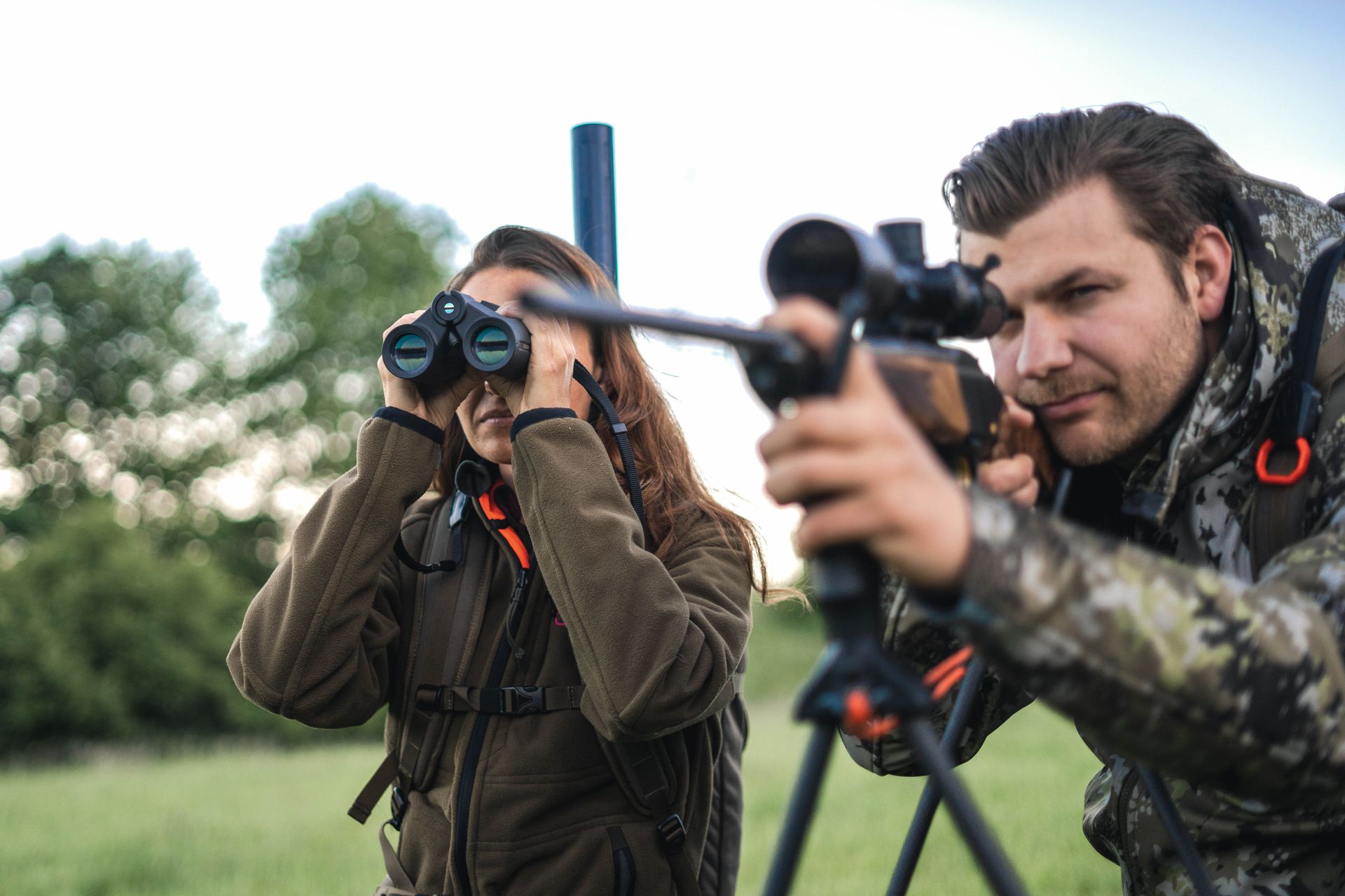 To jegere, en kvinne med kikkert og en mann som sikter med en rifle, på et gresskledd område. De er begge fokuserte og kledd i kamuflasje.