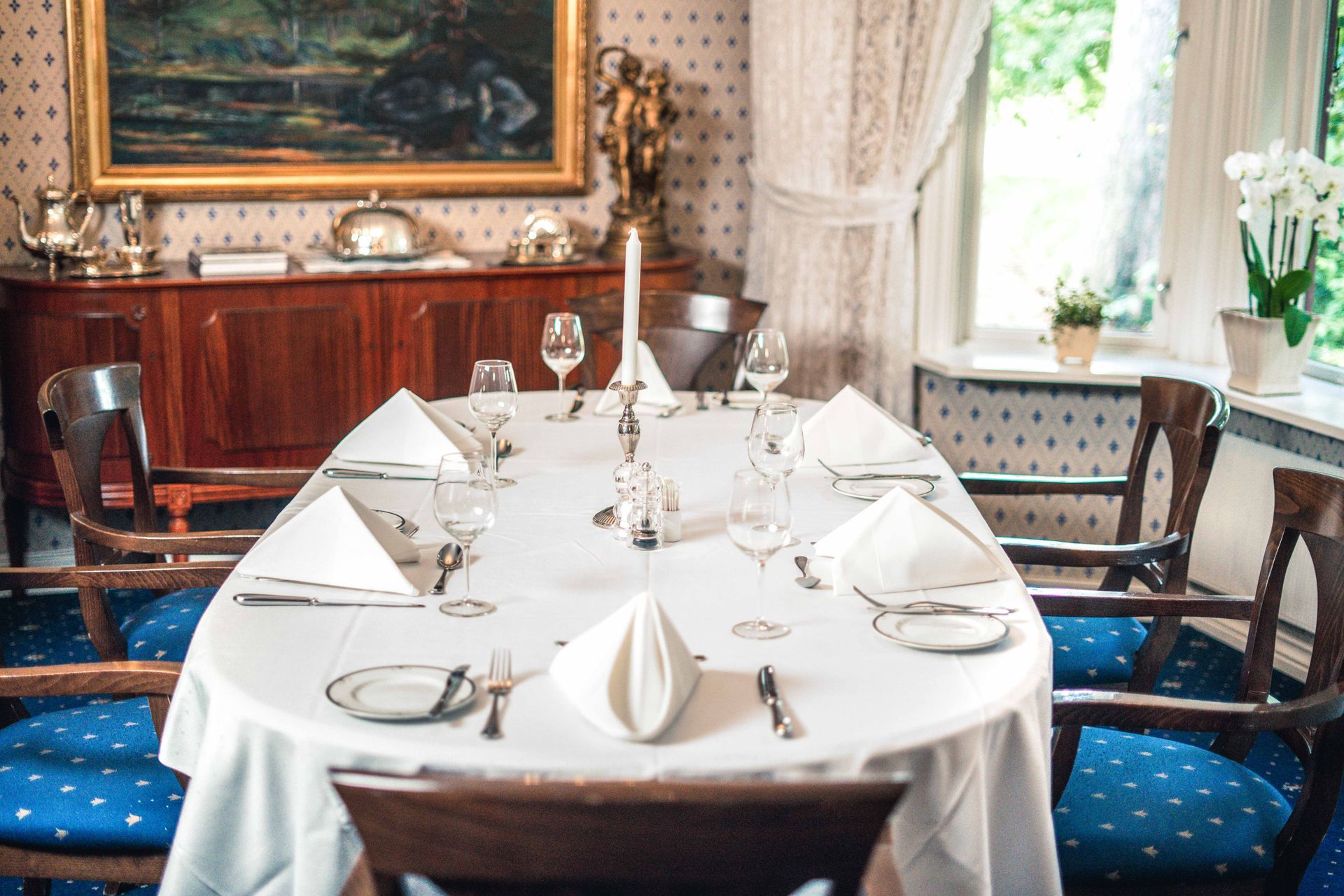 Formelt spisebord dekket til et måltid, med hvitt sengetøy, glass og brettede servietter, i et godt opplyst rom med utsmykkede møbler.