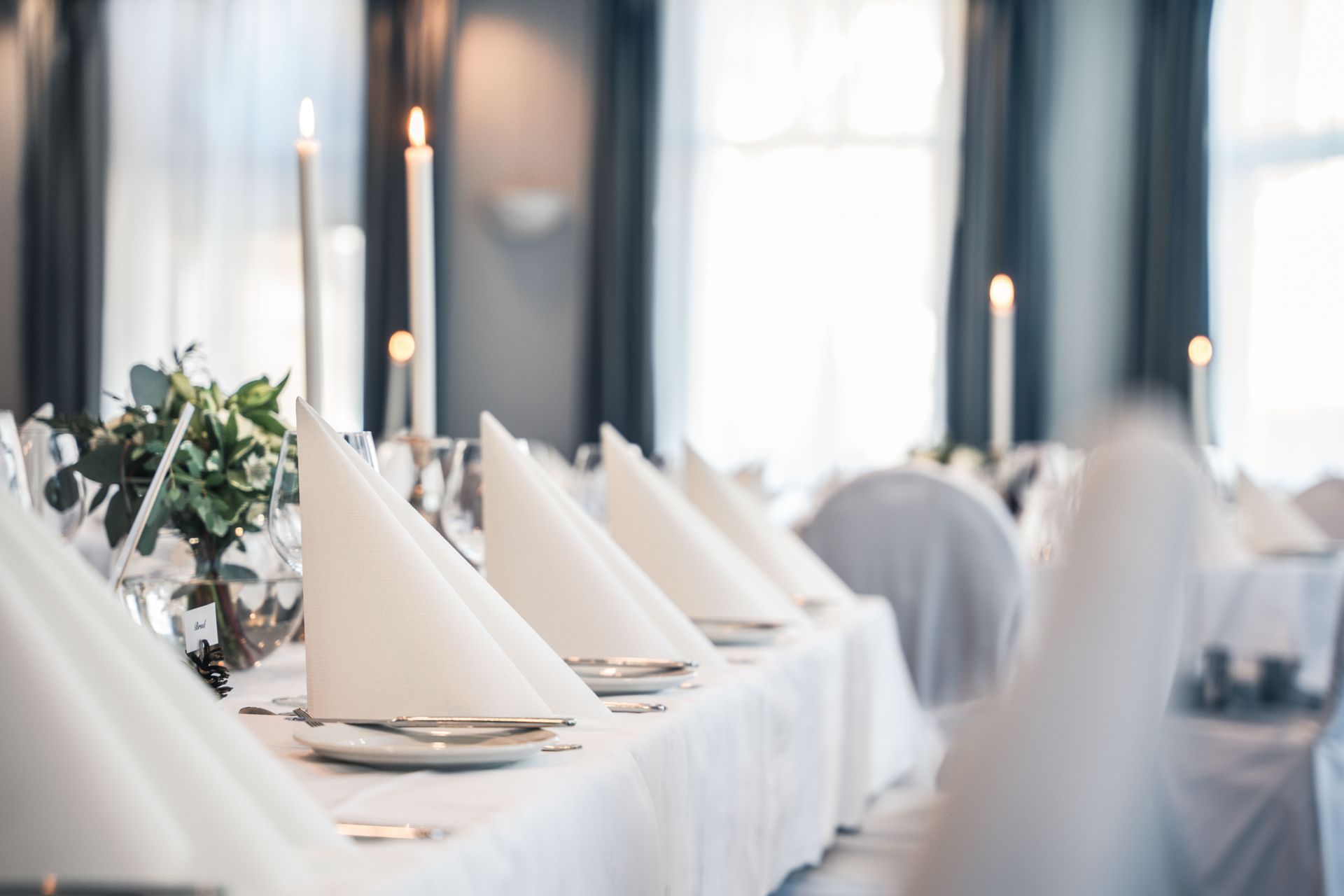 Formelt spisebord dekket til en feiring med hvitt sengetøy, brettede servietter, lys og blomsteroppsatser.