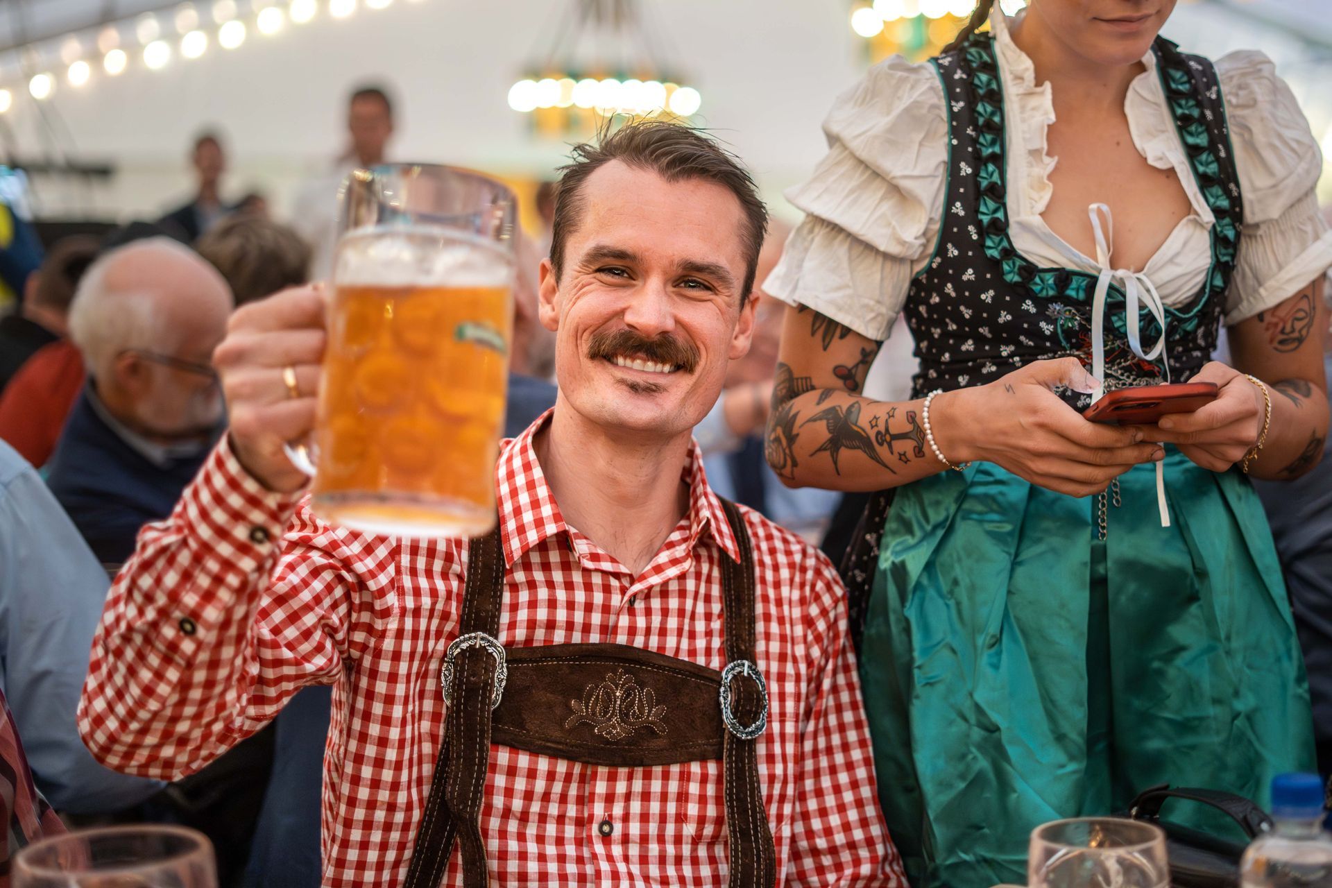 En mann med en stor ølstein skåler på Oktoberfest, ikledd tradisjonelle lederhosen og smilende. En kvinne i dirndl står ved siden av ham og holder en telefon.