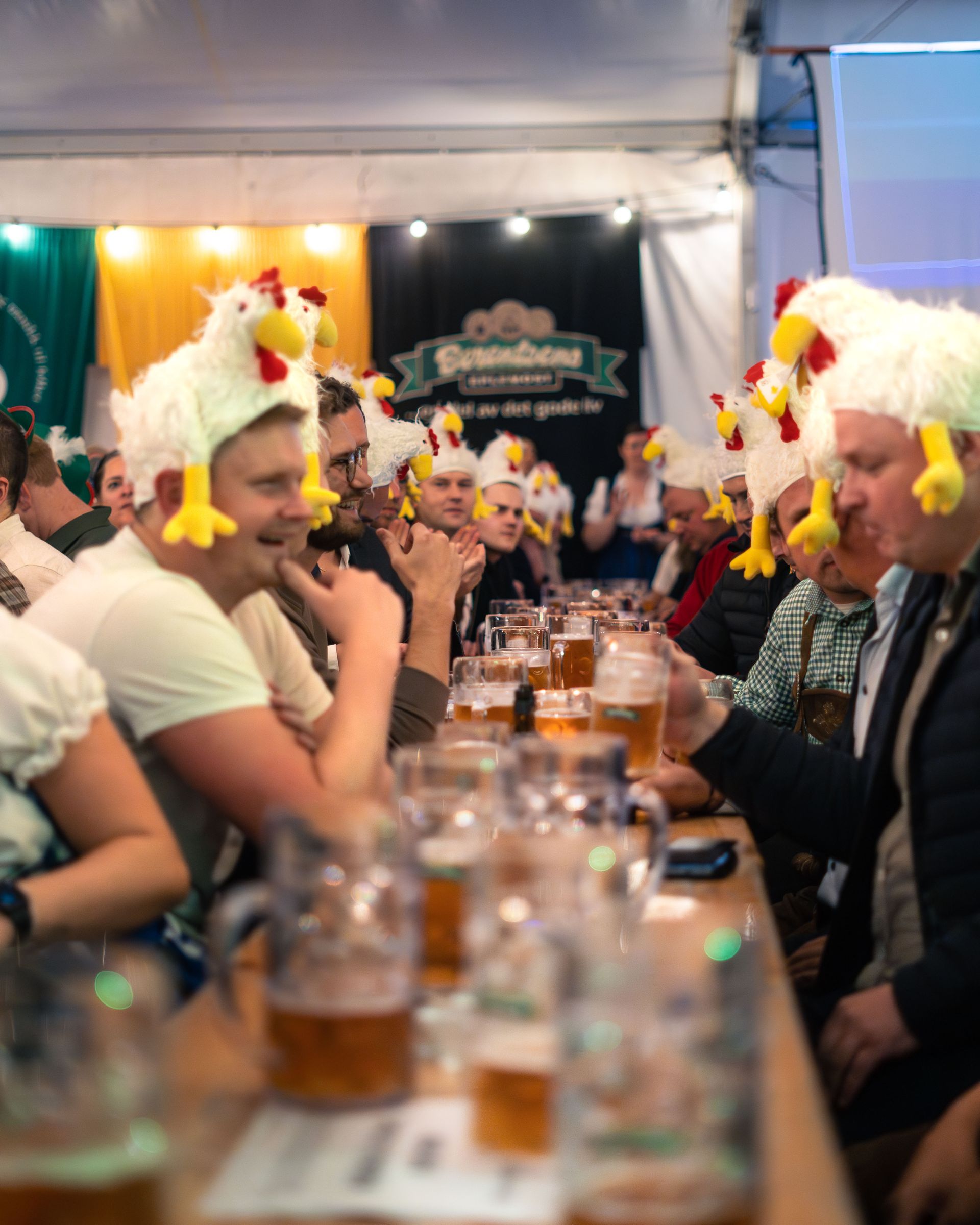 Folk med kyllinghatter drikker øl ved et langt bord i et øltelt. Mange ølkrus er synlige, og settingen er en festival.