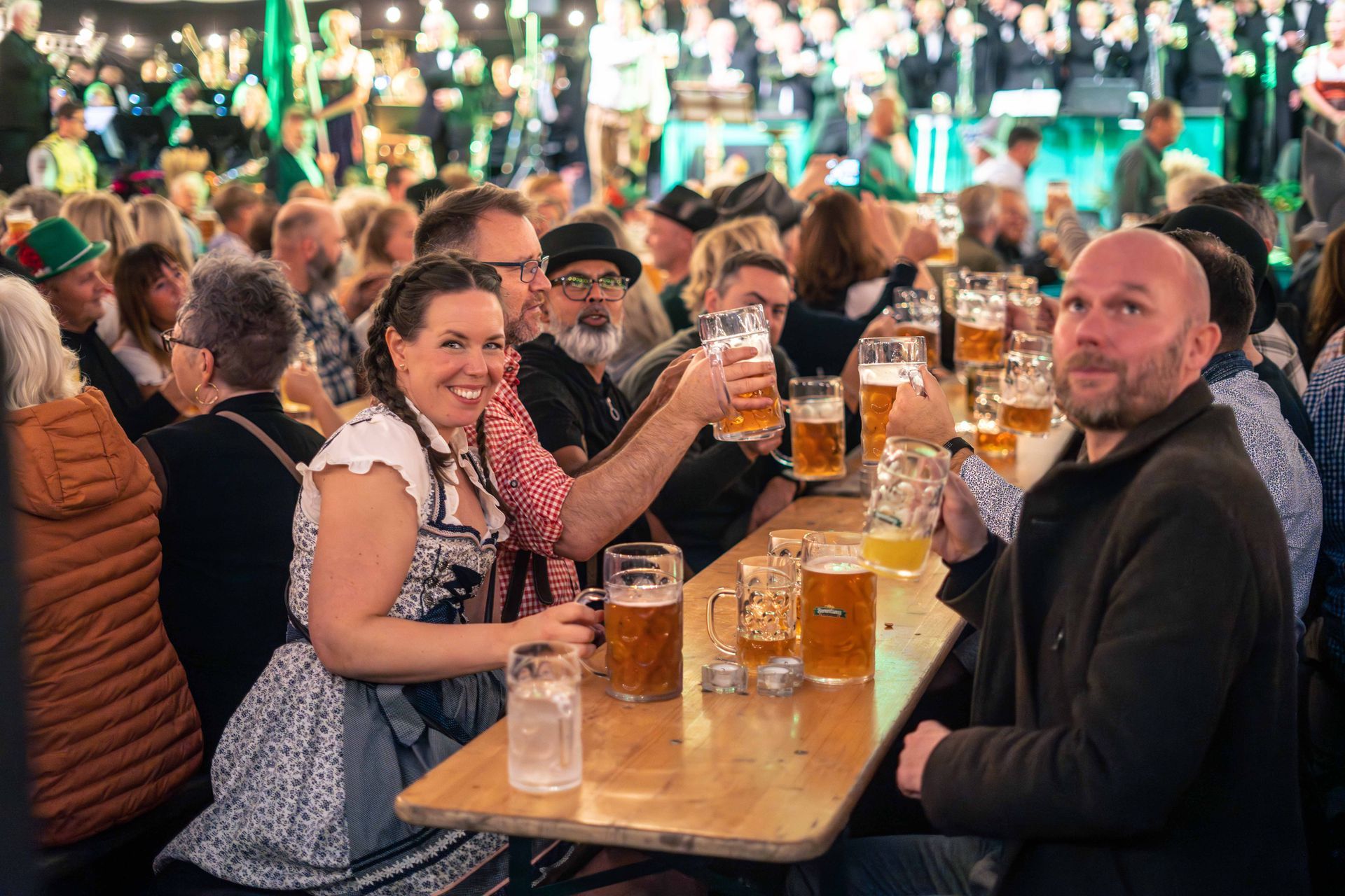 Folk på en festival løfter ølkrus for en skål. En kvinne i dirndl smiler bredt, og mange andre koser seg rundt trebord.