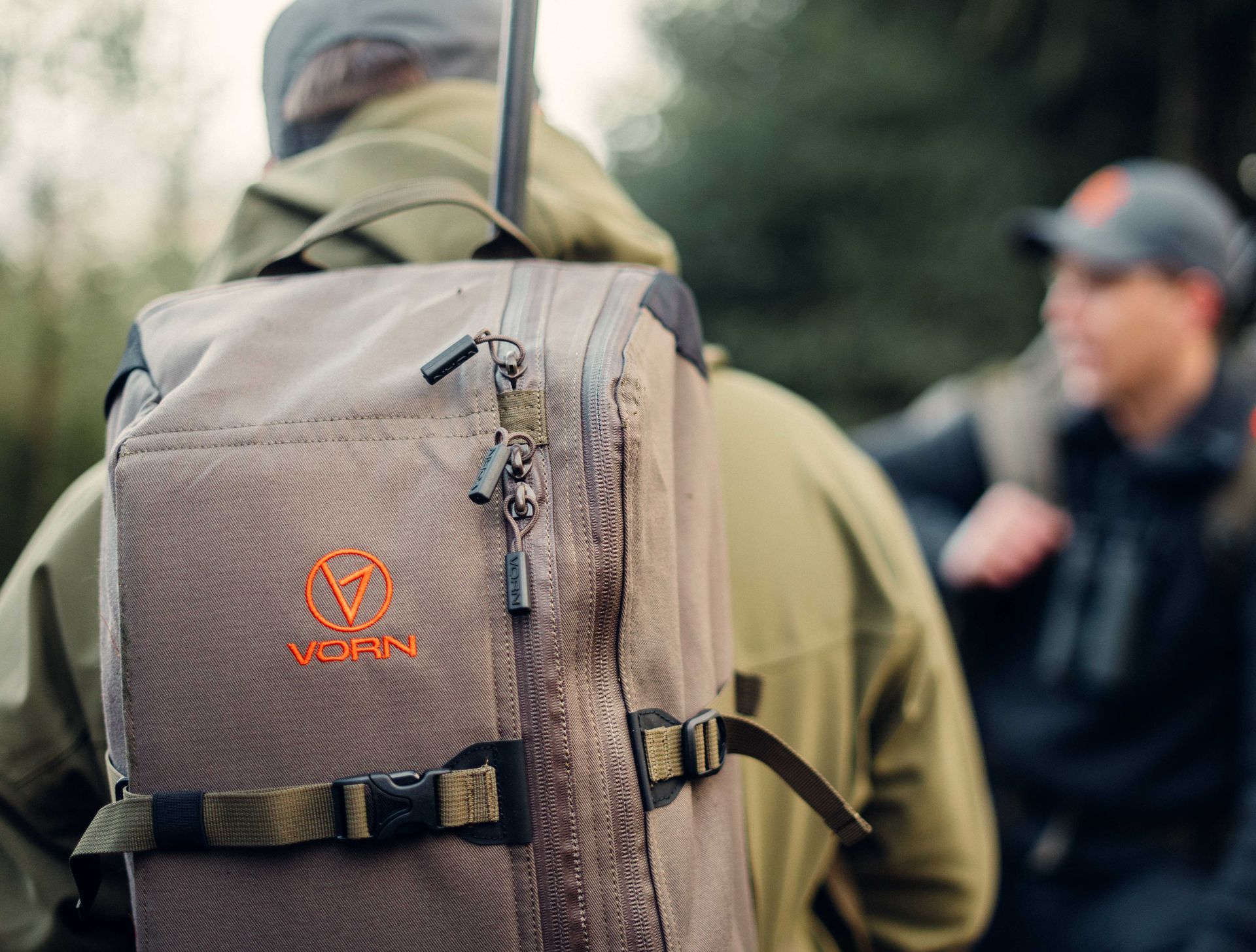 En person i en skog har på seg en brun ryggsekk med en rifle festet til. En annen person er i bakgrunnen.