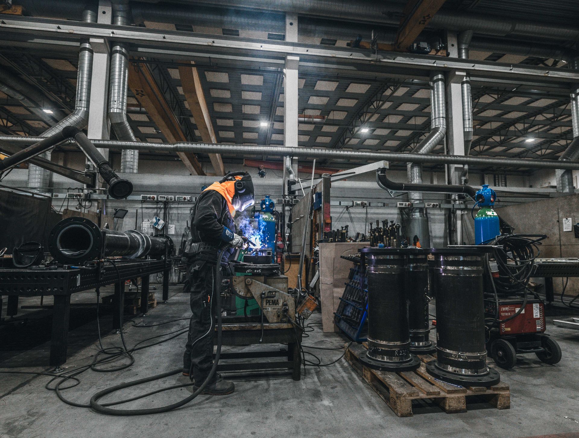 En sveiser med verneutstyr jobber i en fabrikksetting. De sveiser metall i nærheten av flere store, sylindriske gjenstander.