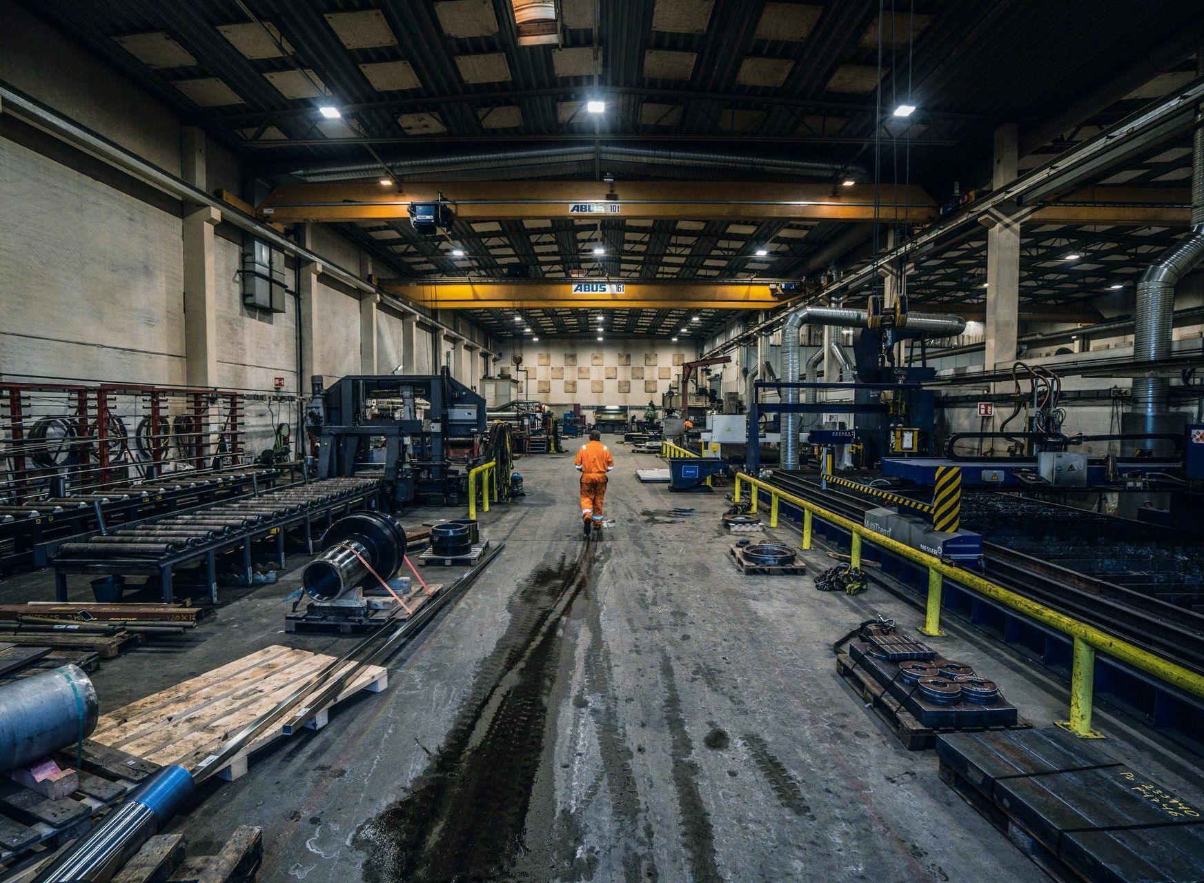 En arbeider i oransje kjeledress går gjennom en stor, industriell fabrikk. Fabrikken er fylt med maskiner, rør og utstyr.