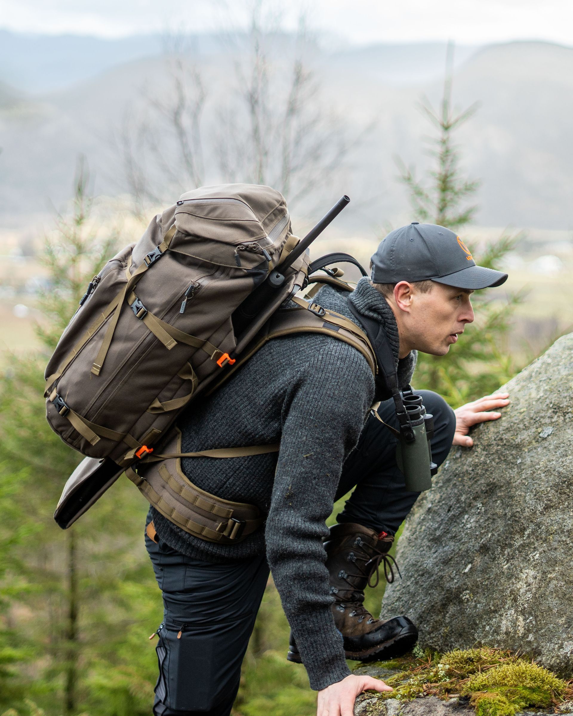 Mann klatrer opp en steinete bakke med en stor ryggsekk og rifle, iført lue og mørk genser. Bakgrunnen viser en fjellkjede.