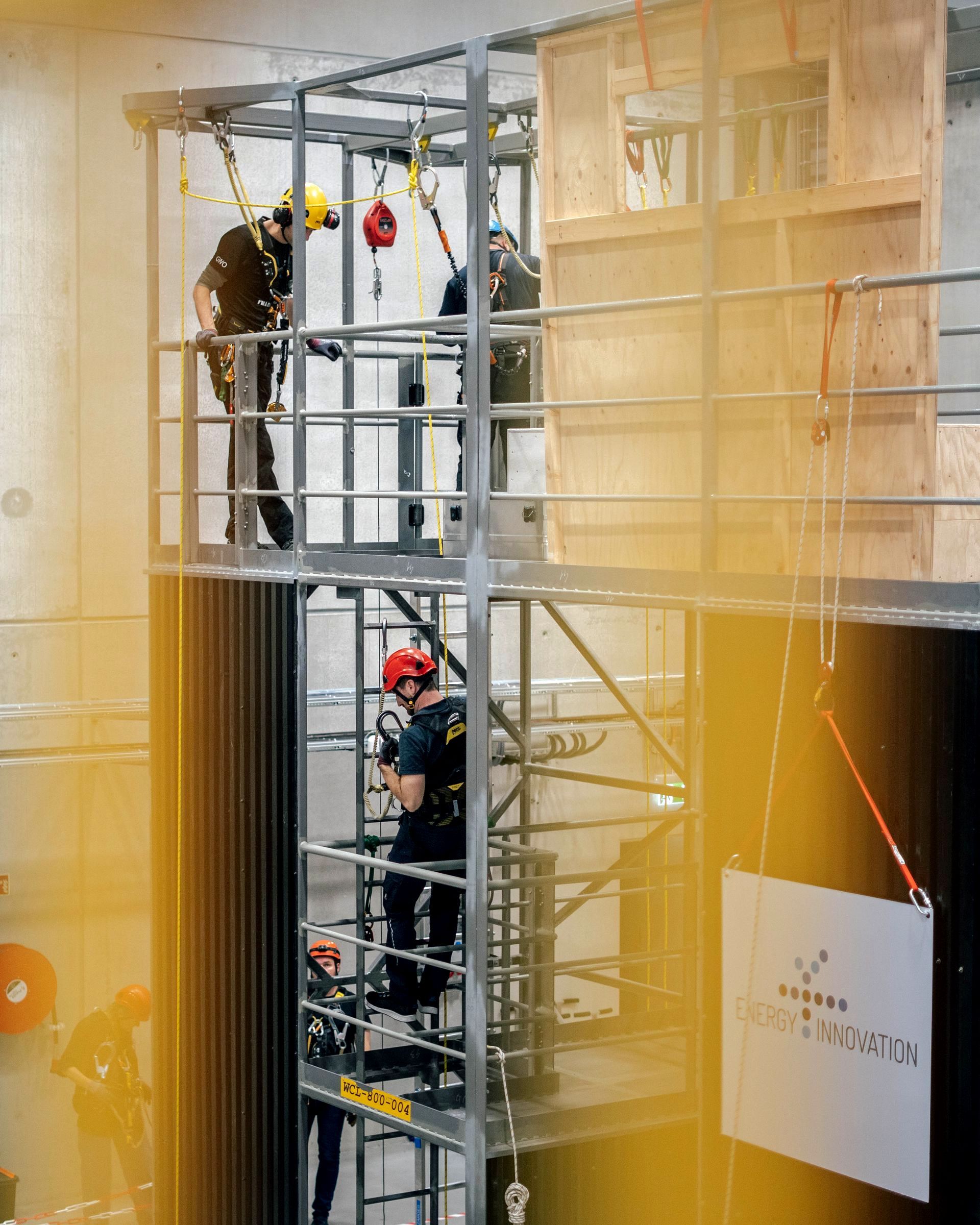 Folk i seler på et metalltreningstårn med trevegg, som øver på redningsteknikker. Innendørs omgivelser, sterk belysning.