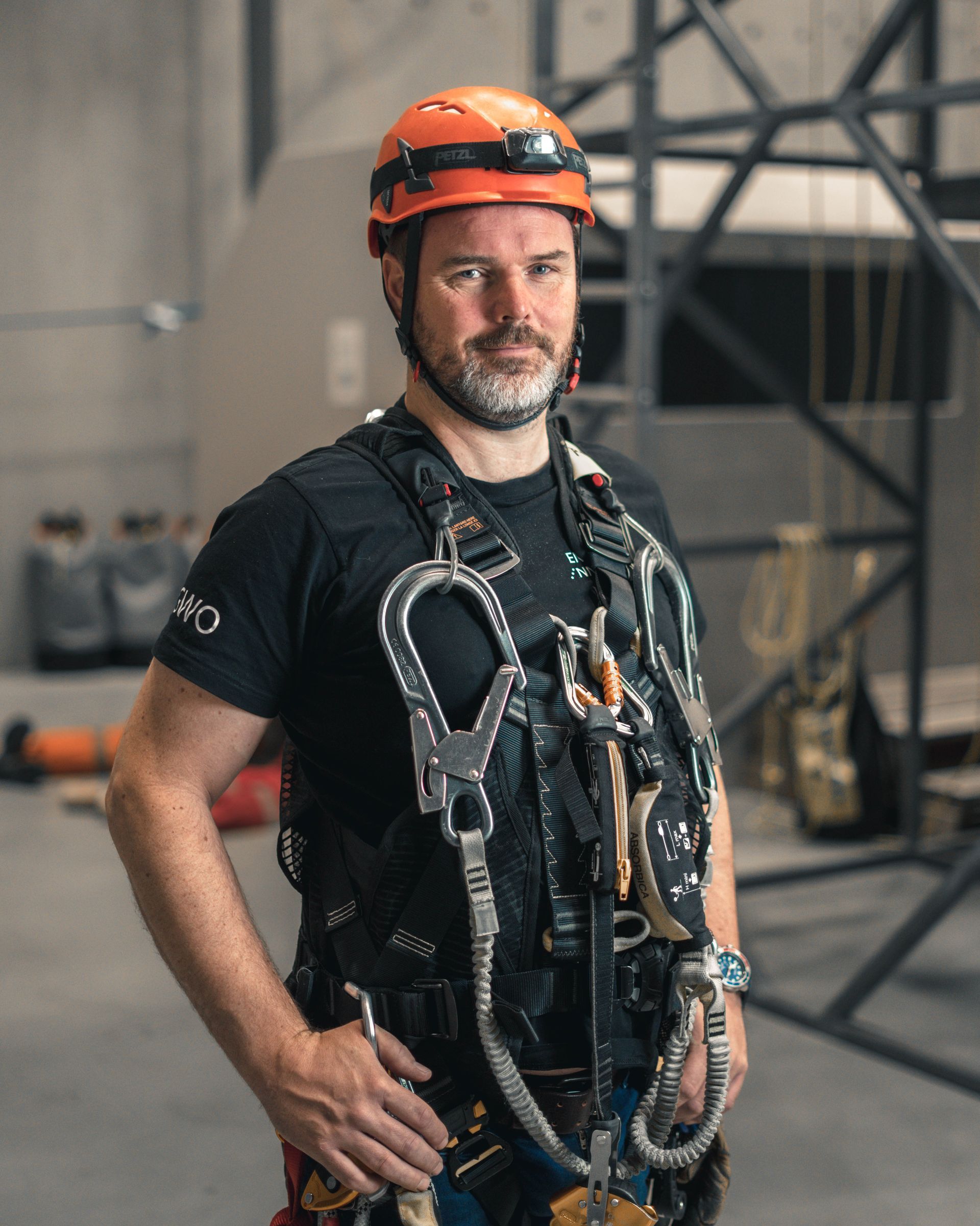 Mann i oransje hjelm og sikkerhetssele poserer innendørs, sannsynligvis et treningsanlegg.
