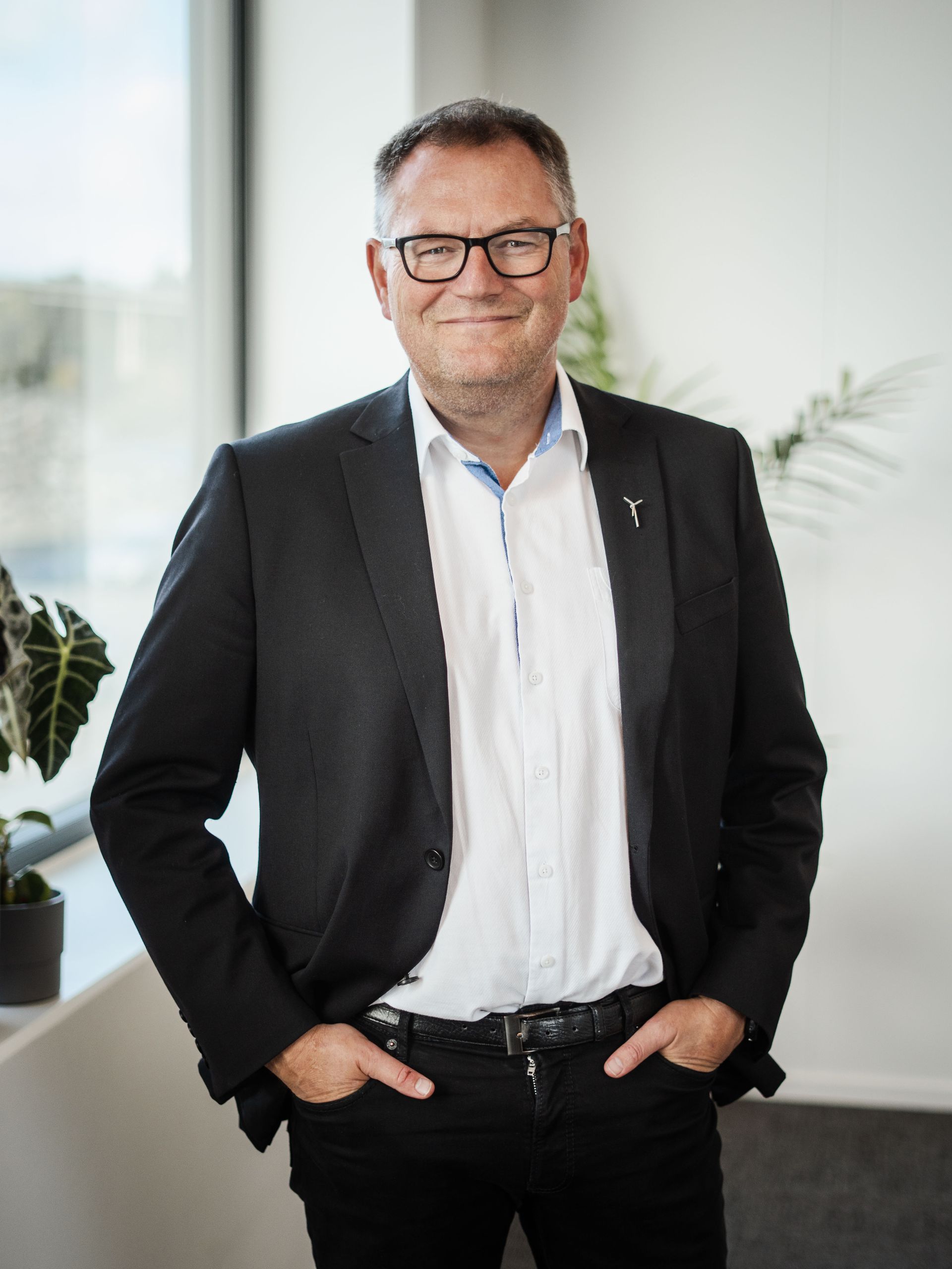 Mann i blazer og briller, hendene i lommene, smilende. Står innendørs ved et vindu og grøntområder.