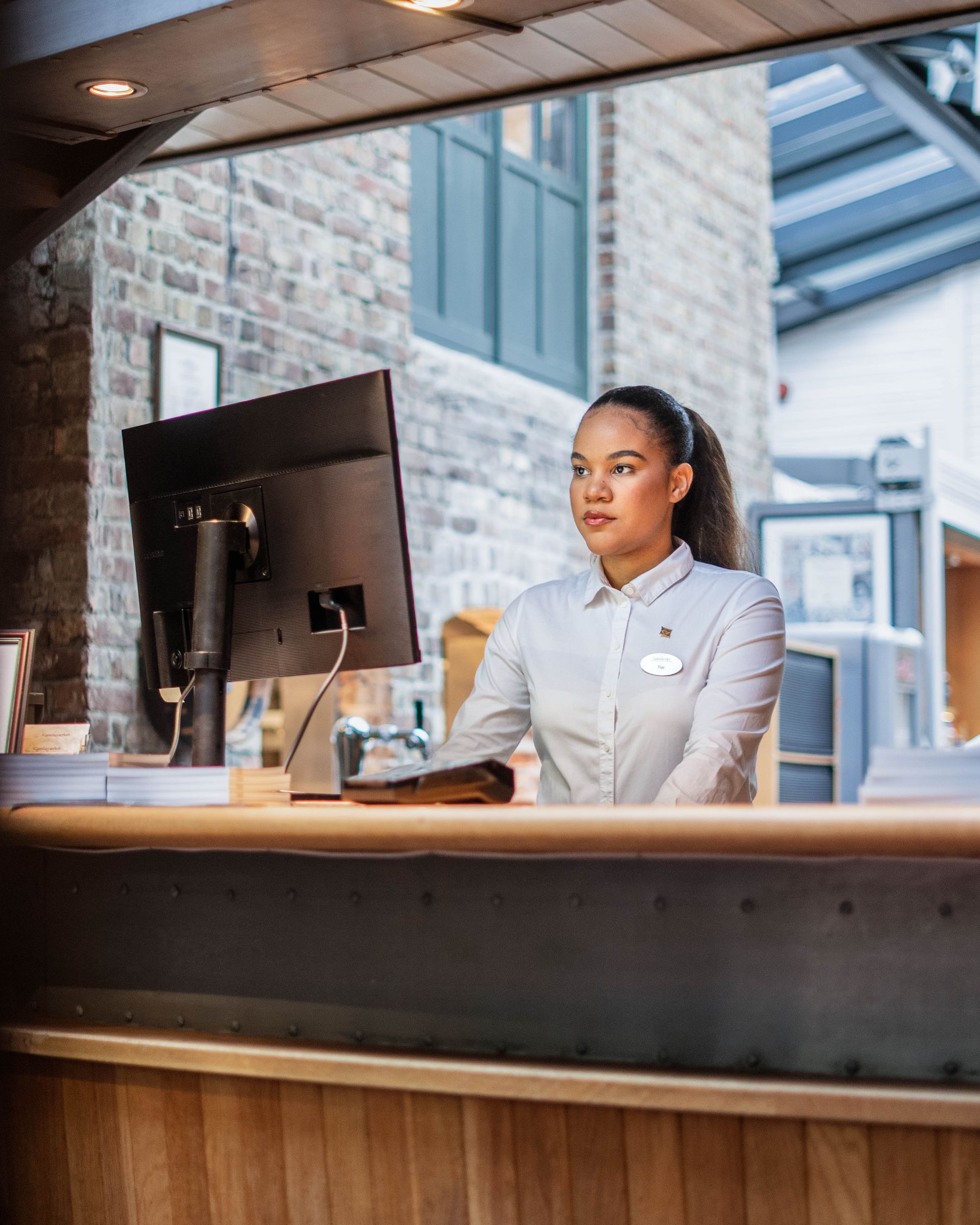 Kvinne i en resepsjonsskranke på et hotell, iført hvit skjorte. Hun ser til høyre. Murvegg i bakgrunnen.
