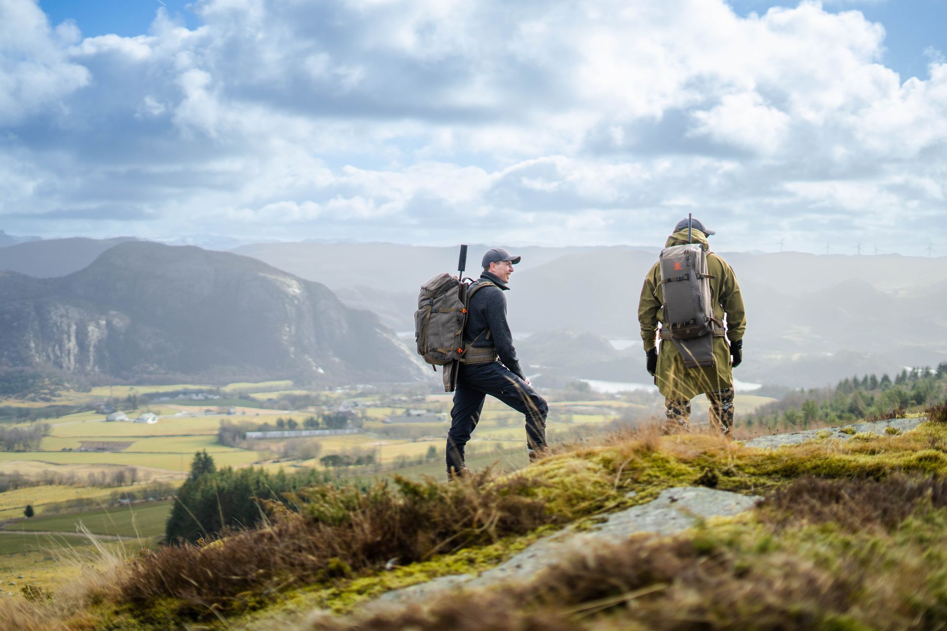 To turgåere med ryggsekker står på en åstopp med utsikt over en dal. Den ene har på seg svart, den andre kamuflasje, på en overskyet dag.