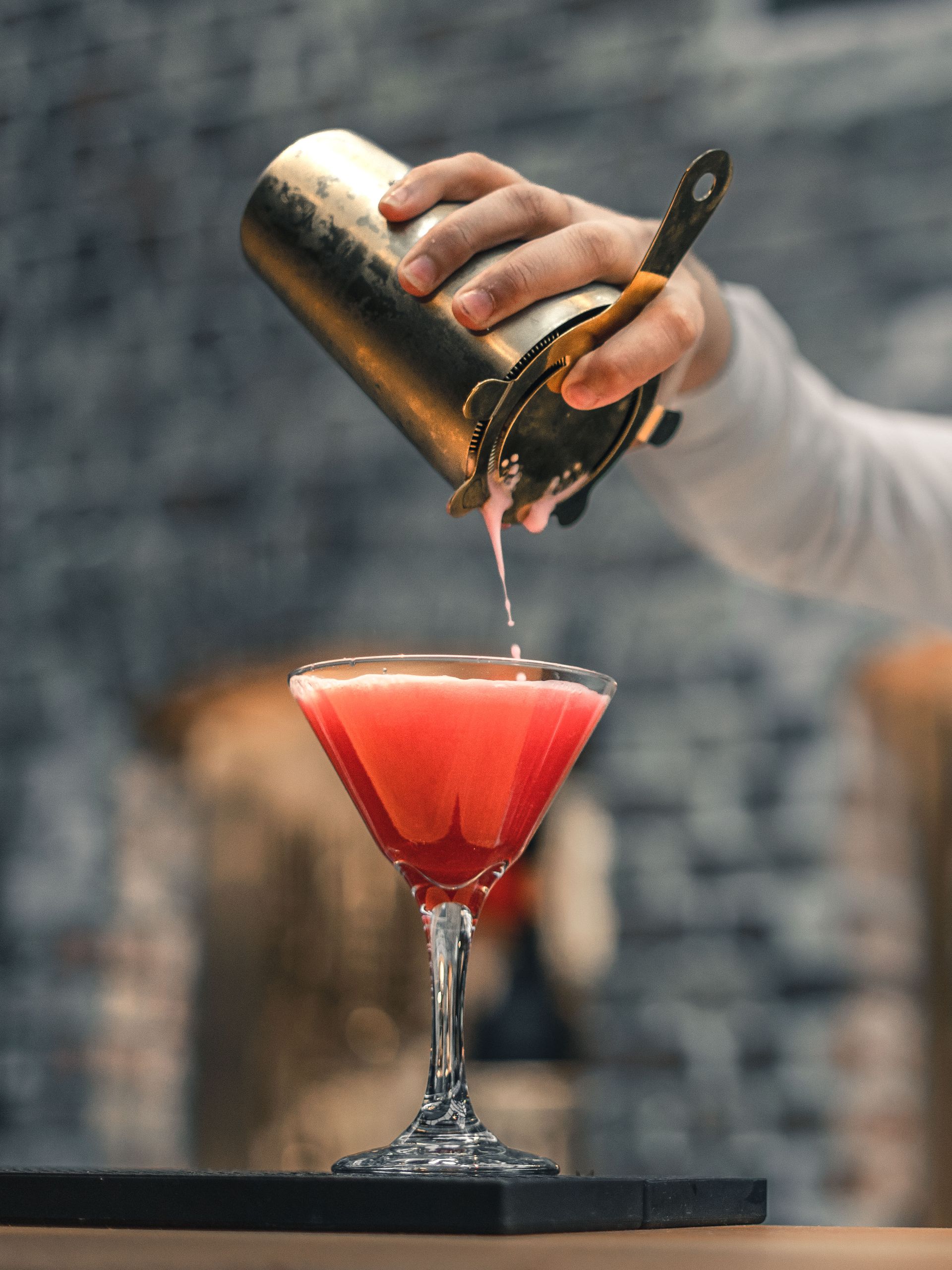 Bartender heller en rosa cocktail fra en gullshaker i et martiniglass. Bakgrunn av en murvegg.