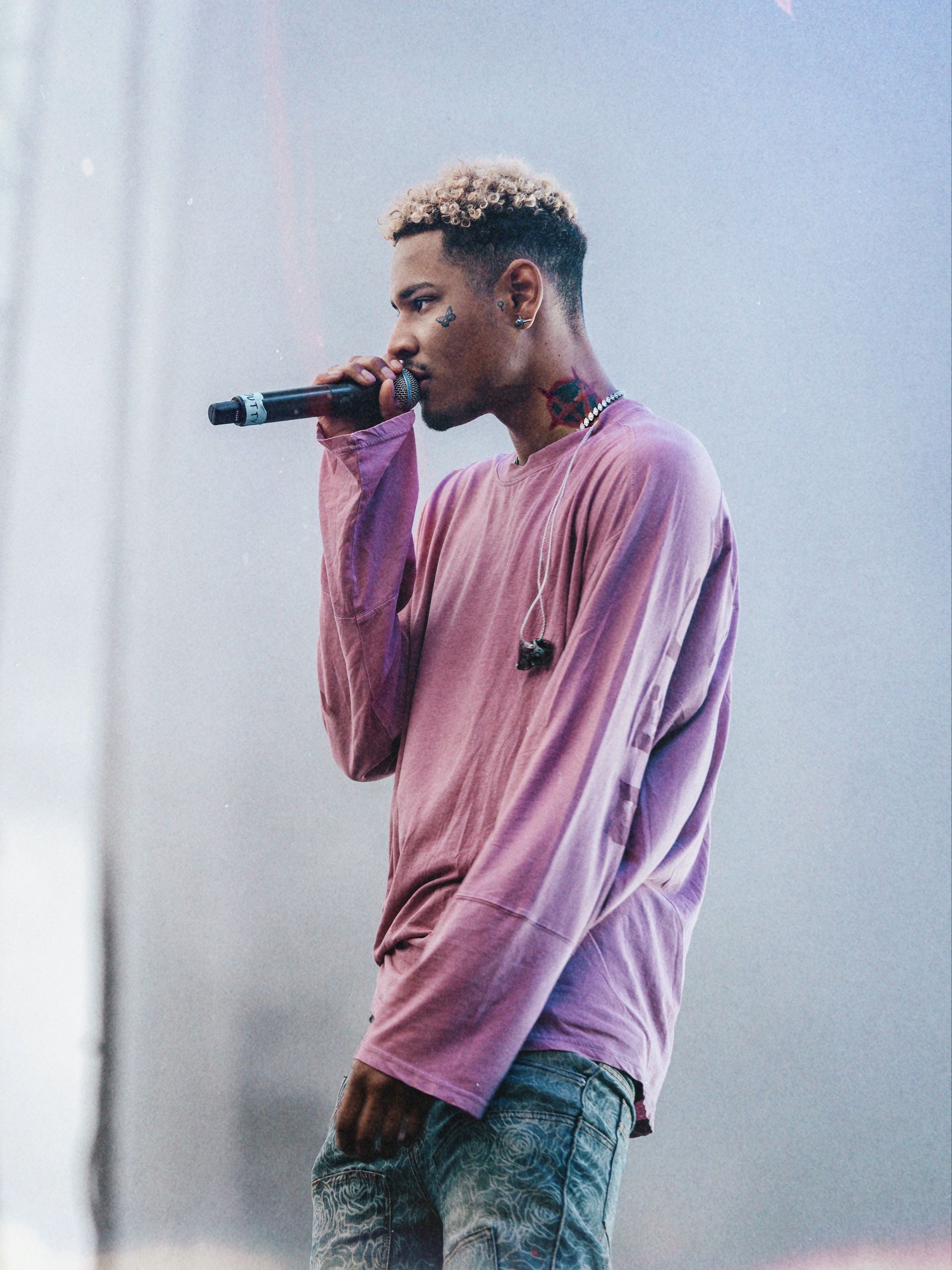 En mann med blondt hår synger inn i en mikrofon. Han har på seg en rosa langermet skjorte og slitte jeans, mens han opptrer på scenen.