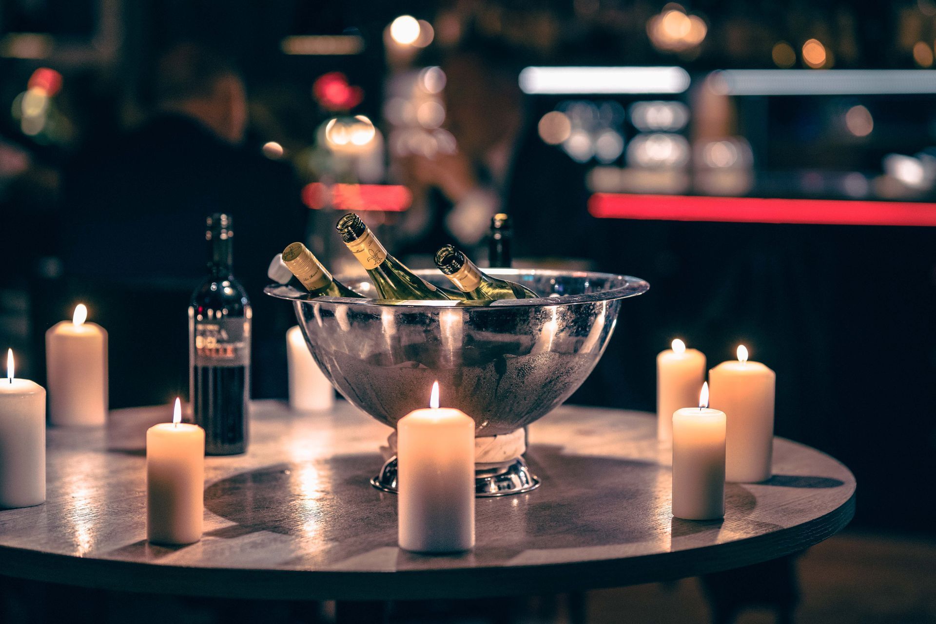 Rundt bord med stearinlys og en sølvfarget vinkjøler, vinflasker, i en svakt opplyst restaurantsetting.