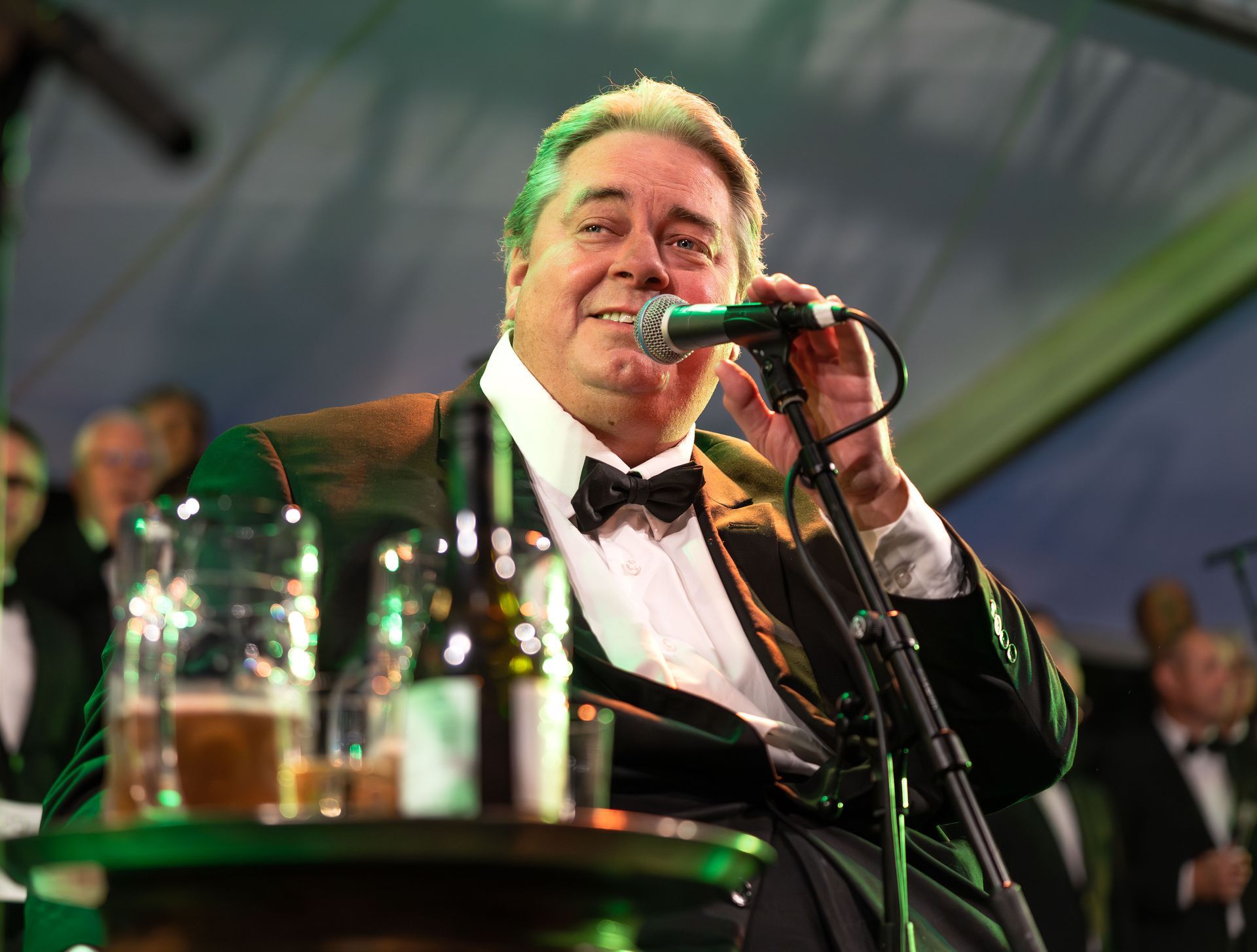 Mann i smoking synger inn i en mikrofon, holder den med én hånd, smiler. Drinker foran seg, band i bakgrunnen.