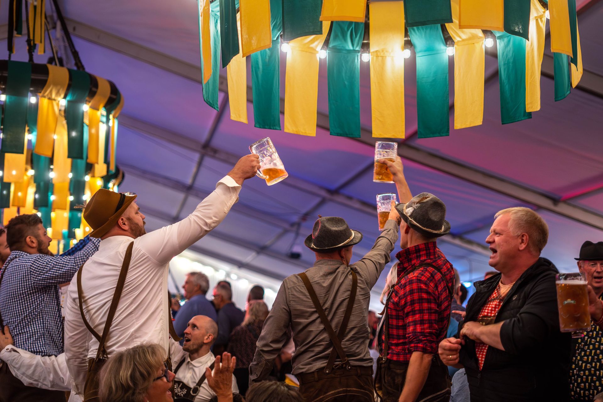 Folk i tradisjonelle klær hever ølsteiner i et telt dekorert med gule og grønne serpentiner.