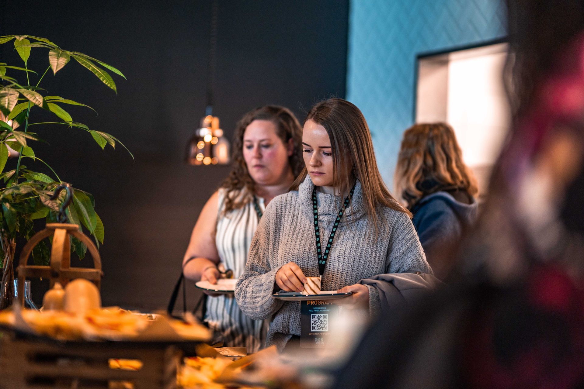 To kvinner ved et buffetbord, den ene i en grå genser, den andre i en stripete topp, tar mat. Mørkt miljø, med planter og lysarmaturer.