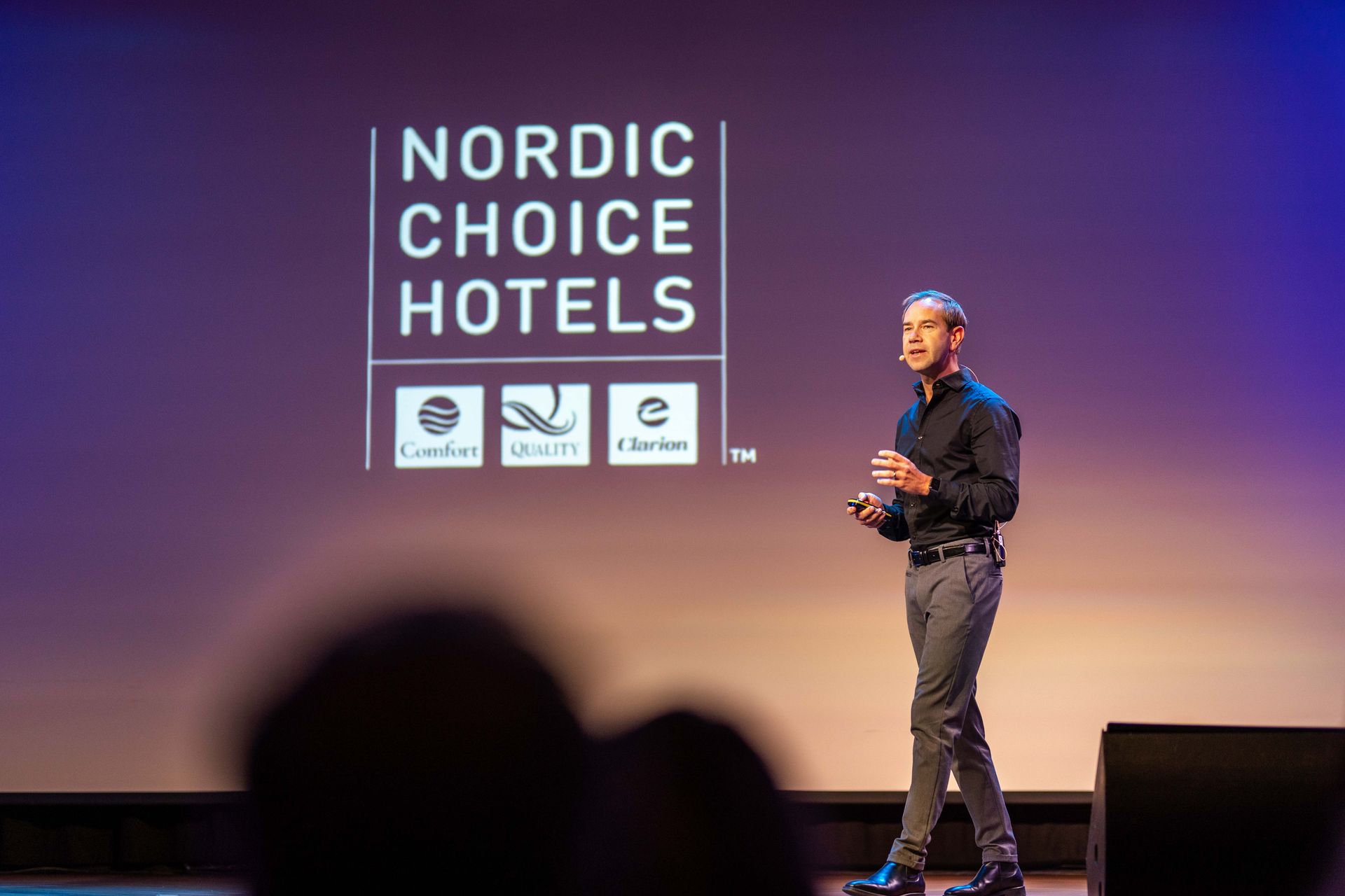 Mann som presenterer på scenen foran en «Nordic Choice Hotels»-logo. Han har på seg en svart skjorte og grå bukser, og snakker inn i en mikrofon.