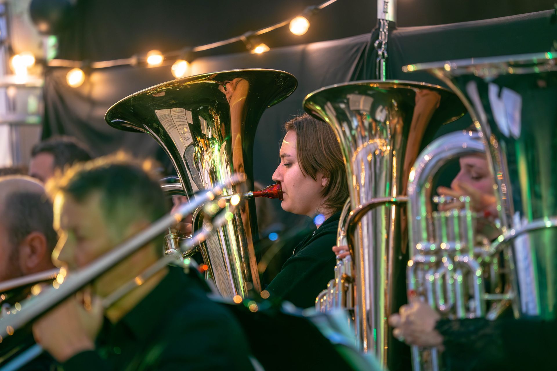 Et band opptrer utendørs, med fokus på tubaister. En kvinne blåser i tubaen, ser fokusert ut, med lyslenker i bakgrunnen.