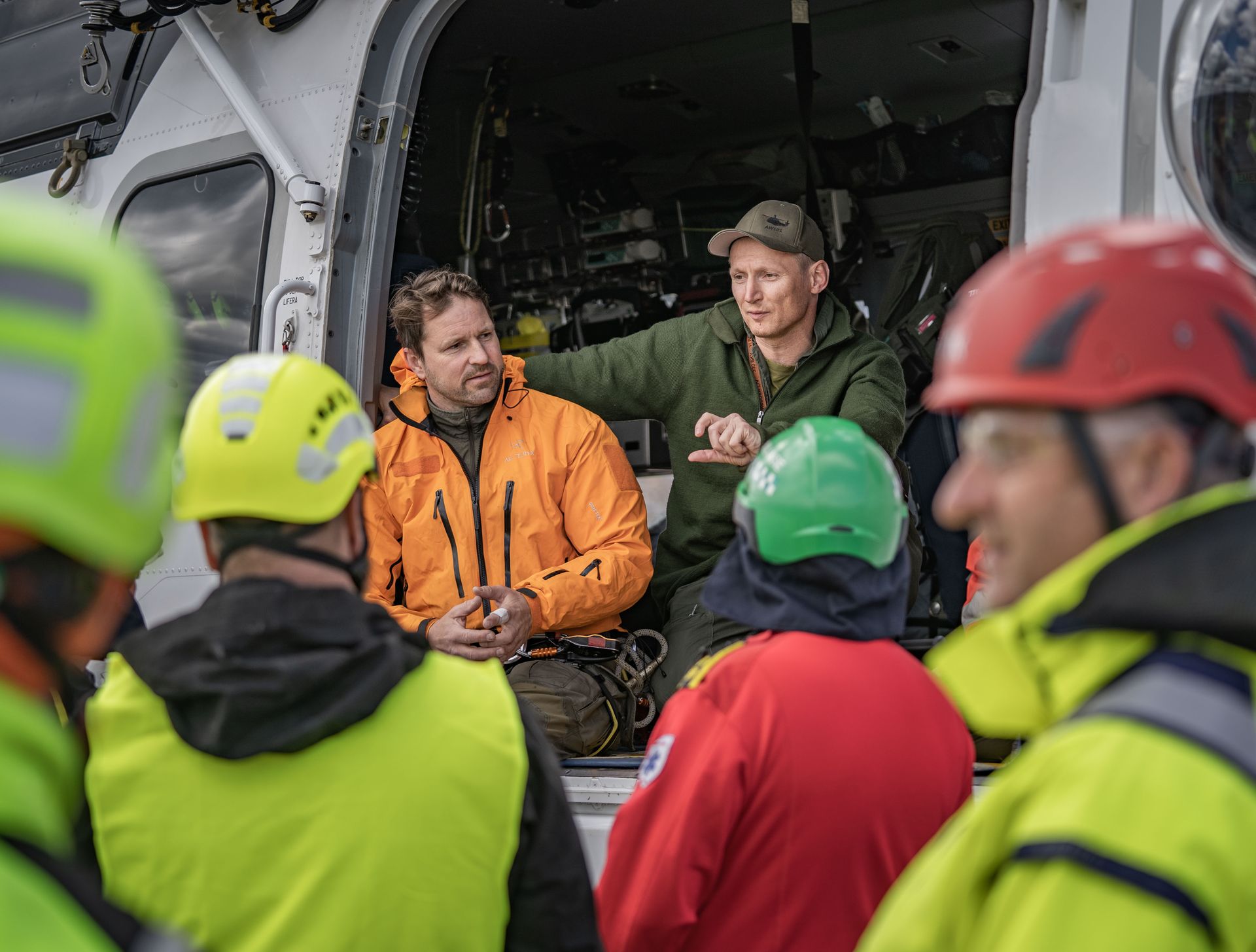 En gruppe mennesker i sikkerhetsutstyr, inkludert hjelmer og jakker, er samlet nær en åpen helikopterdør. En mann i oransje jakke og en annen i grønn uniform snakker inne i helikopteret.