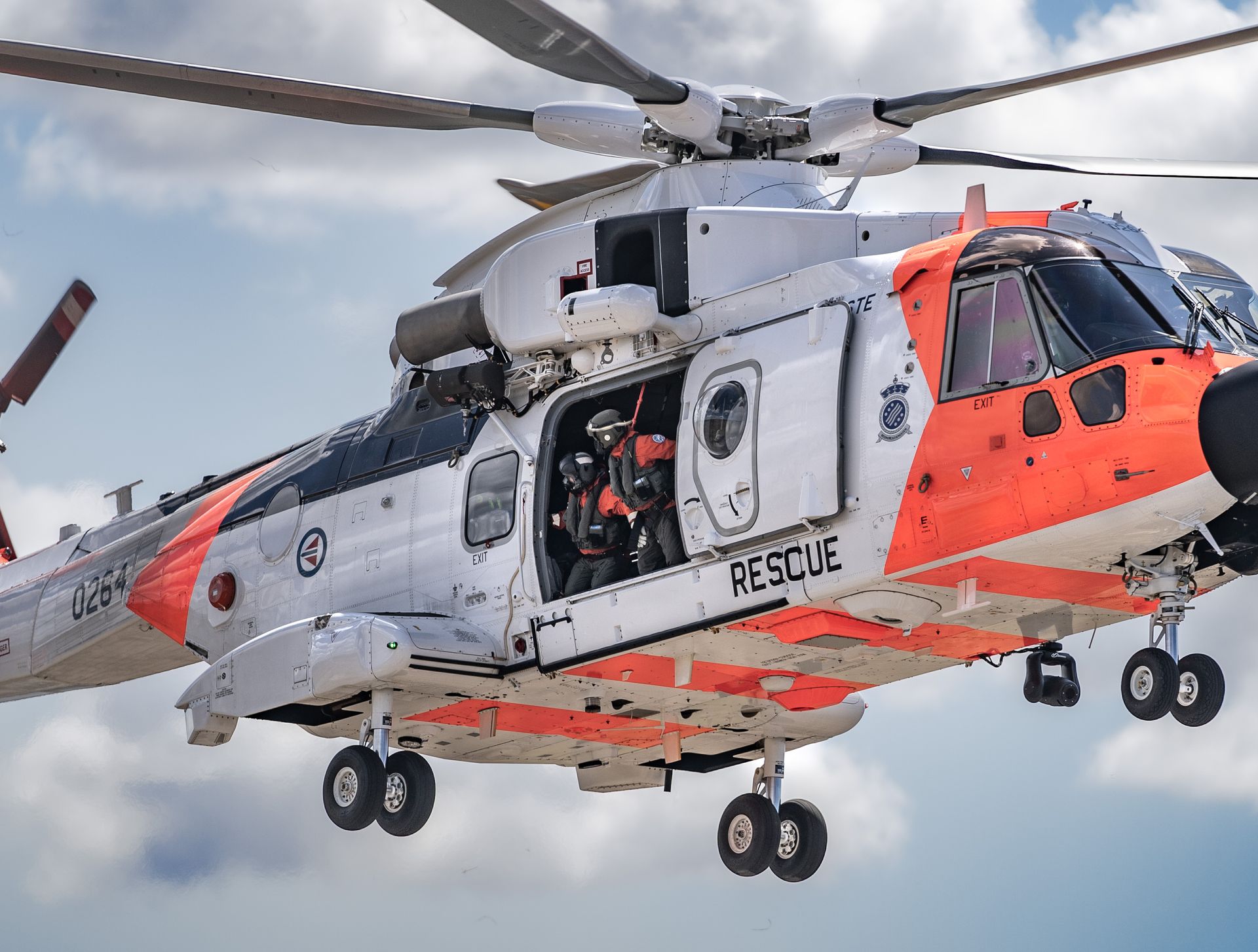 Et hvitt og oransje kystvakthelikopter med åpen sidedør, merket «RESCUE», i luften mot en overskyet himmel.