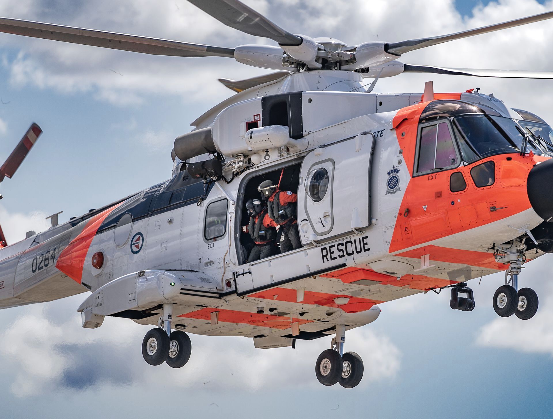 Kystvakthelikopter i luften, hvitt og oransje, med «RESCUE» synlig på siden, mannskap synlig i åpen døråpning.