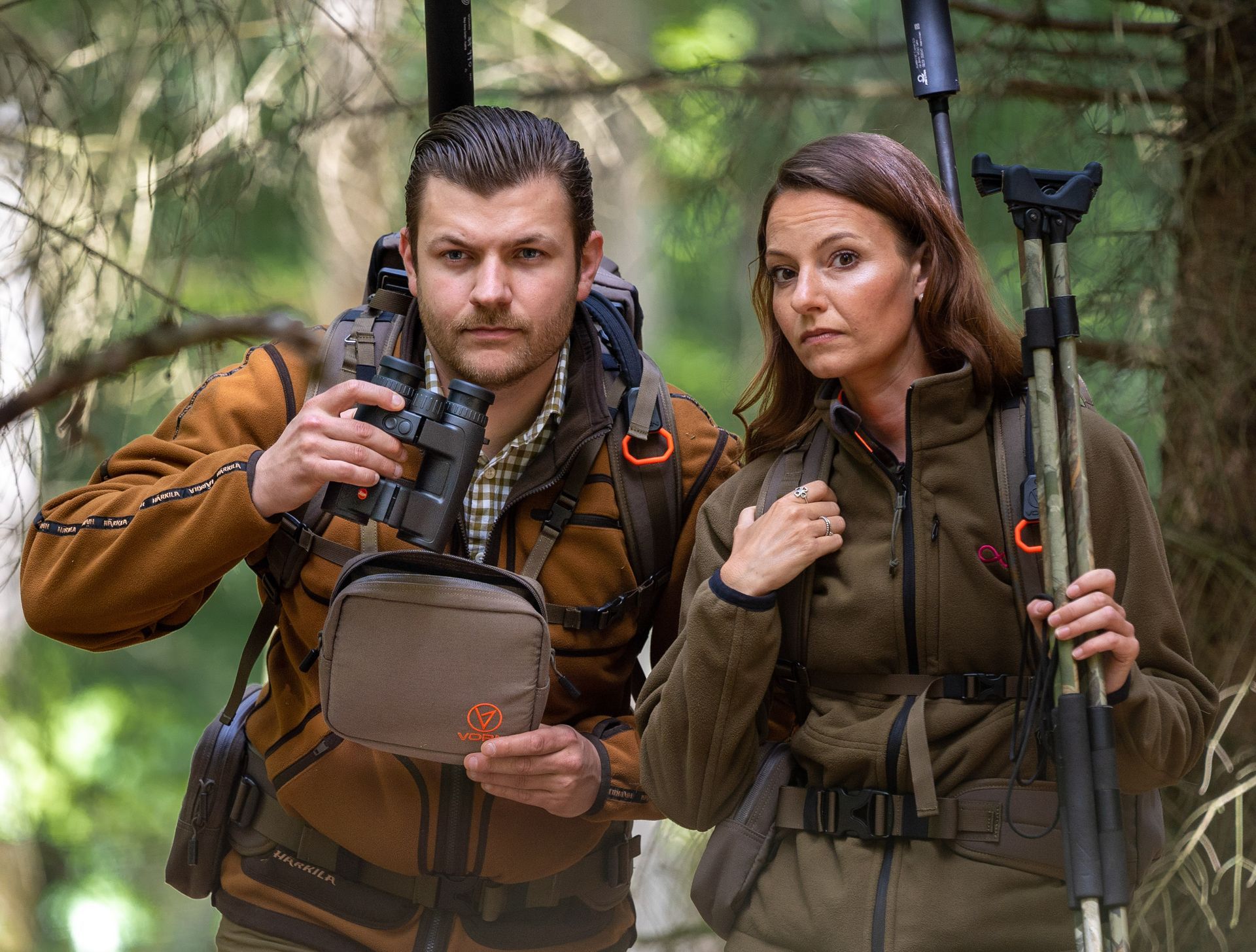 En mann og en kvinne i jaktutstyr står i en skog. Mannen holder en kikkert, kvinnen holder et stativ, begge ser fremover.