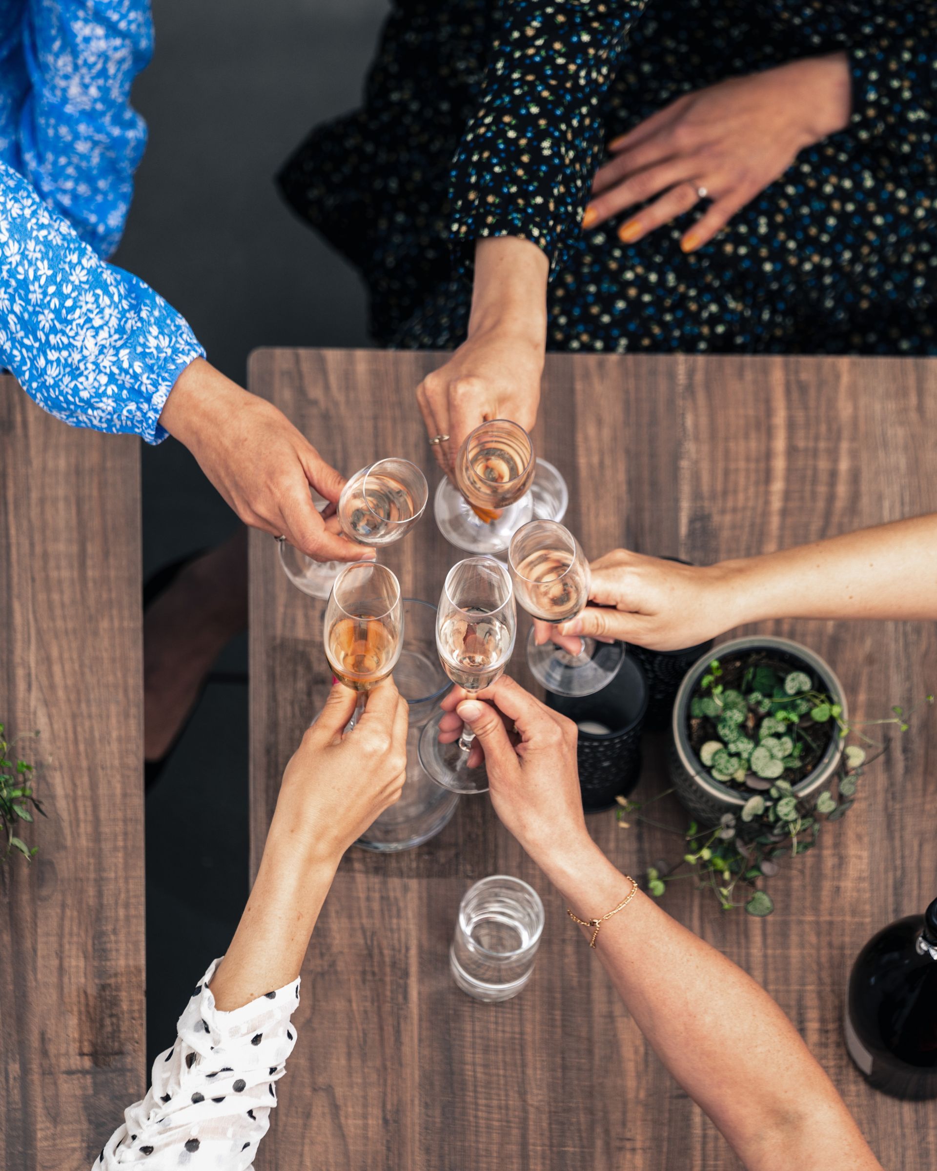Folk skåler med champagneglass rundt et trebord og feirer en sammenkomst. Ulike armer og antrekk er synlige.