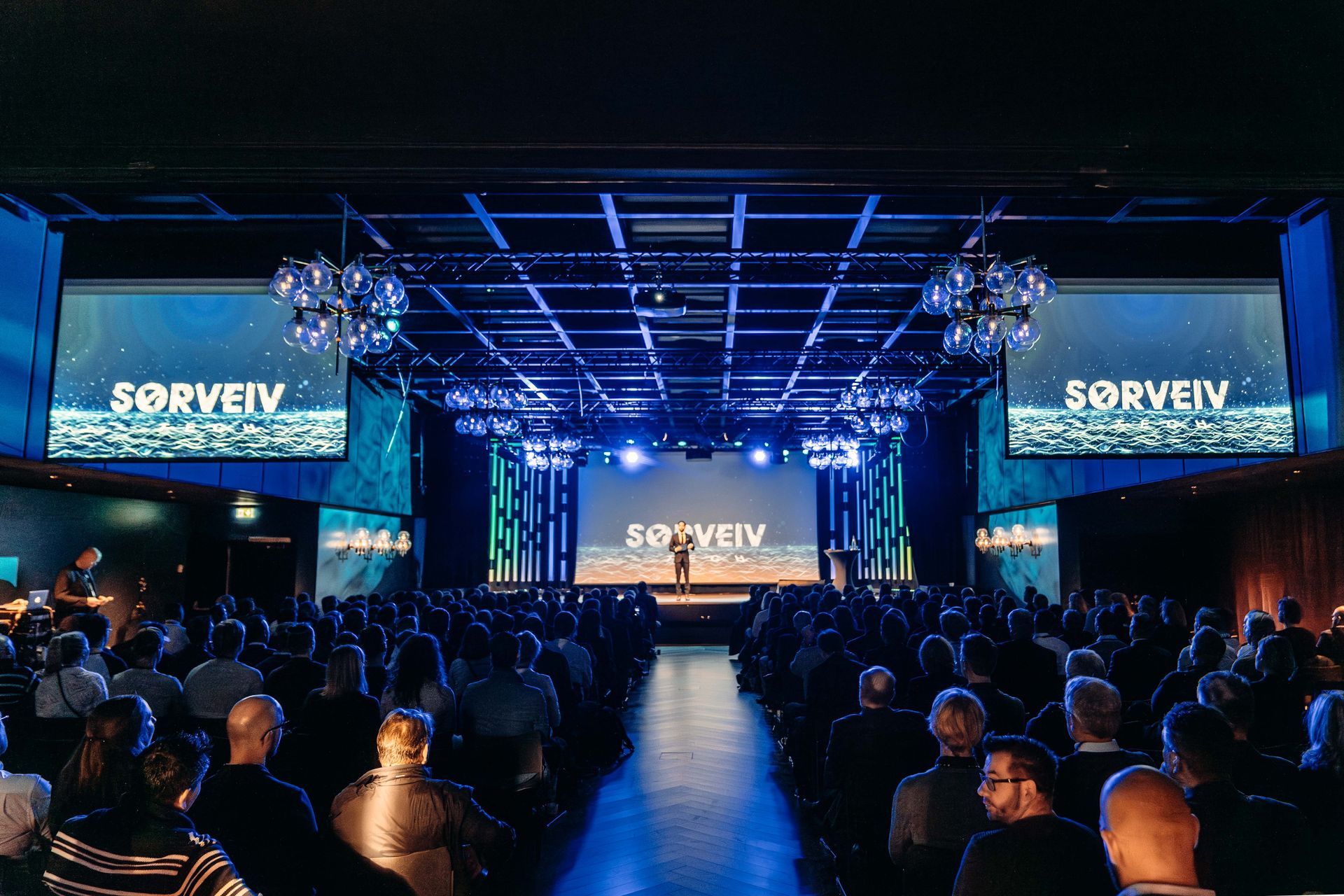 En stor konferansesal med en scene opplyst i blått og med «SORVEIV»-logoen, og et publikum sittende på rekke og rad.