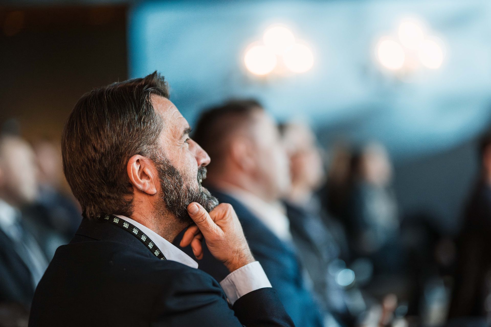 En mann i dress med skjegg berører tankefullt haken sin mens han ser oppover. Han er i en konferansesetting.