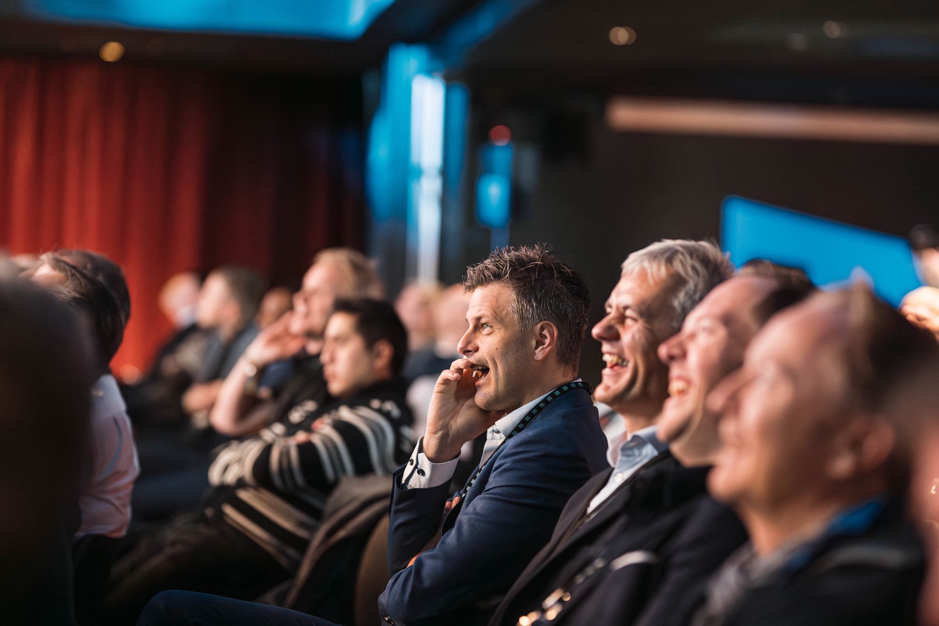 Publikum i et mørkt auditorium ler og lytter. Flere menn er synlige, og én av dem snakker i telefonen.