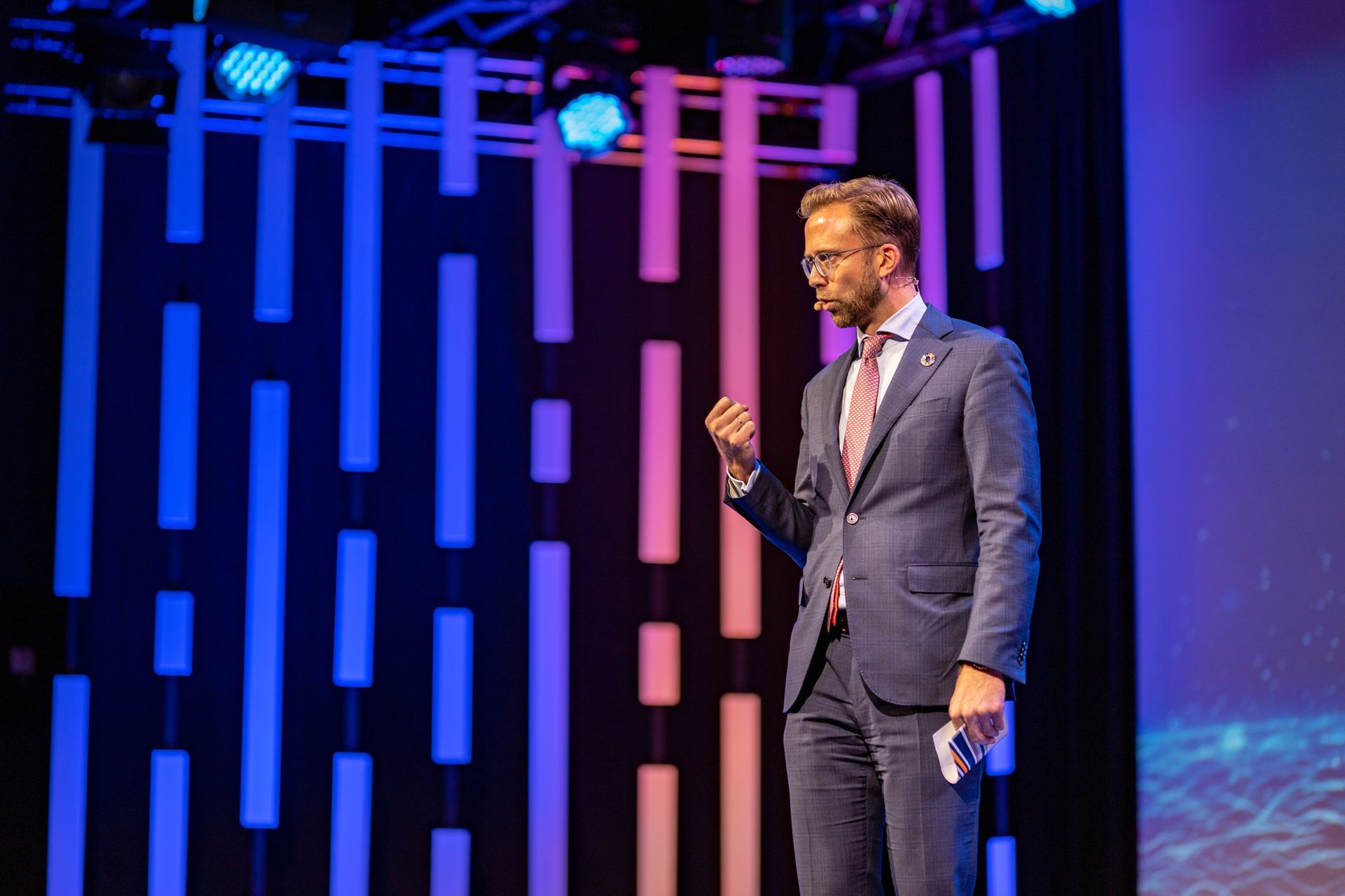 En mann i dress snakker på scenen. Han gestikulerer med høyre hånd mens han holder et lite papir i venstre. Bakgrunnen har vertikale, glødende lys.
