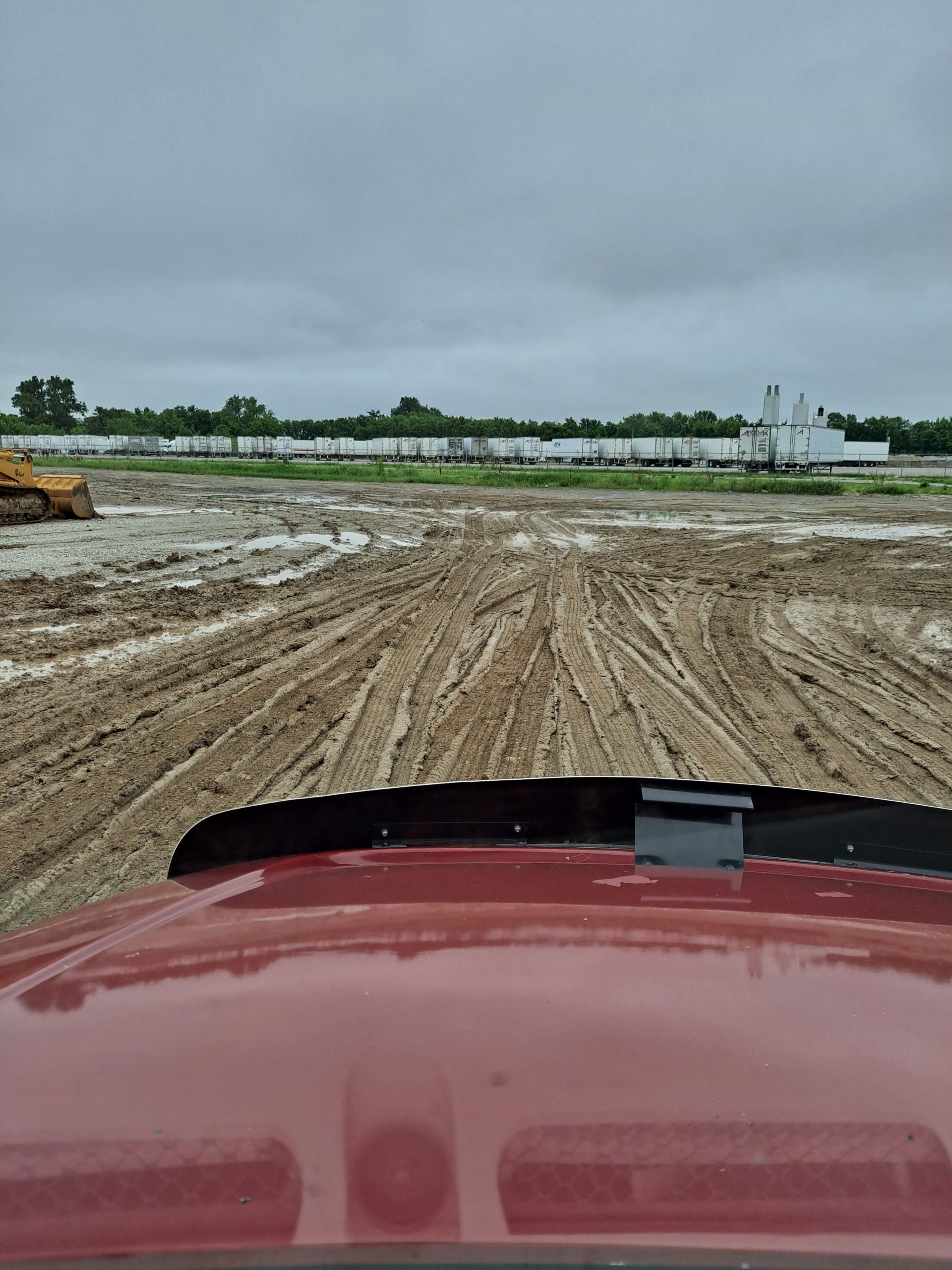 Red vehicle driving through muddy construction site. White buildings in the distance. Overcast sky.