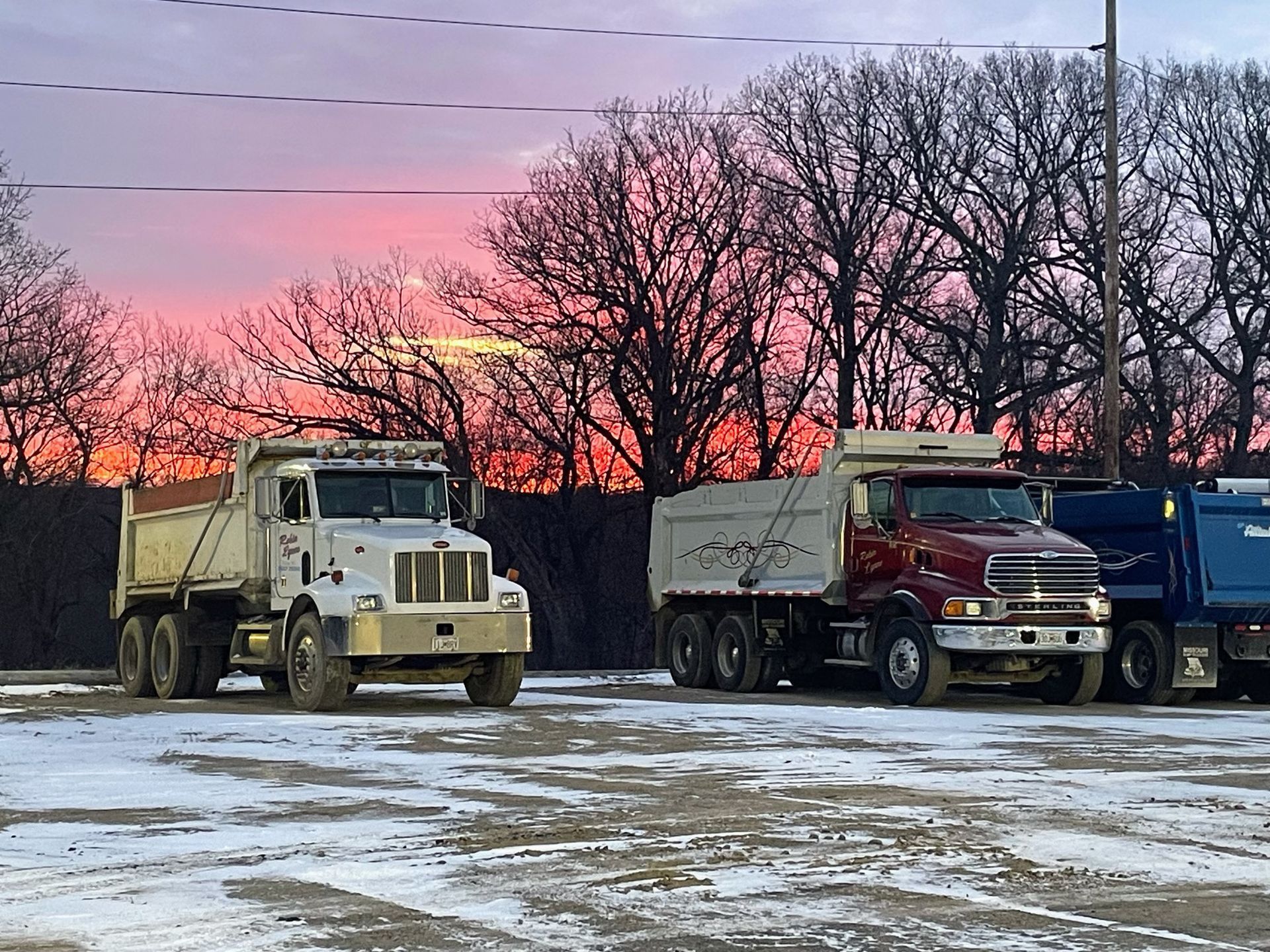 Dump trucks parked in a snowy lot at sunset. The sky is pink and orange.