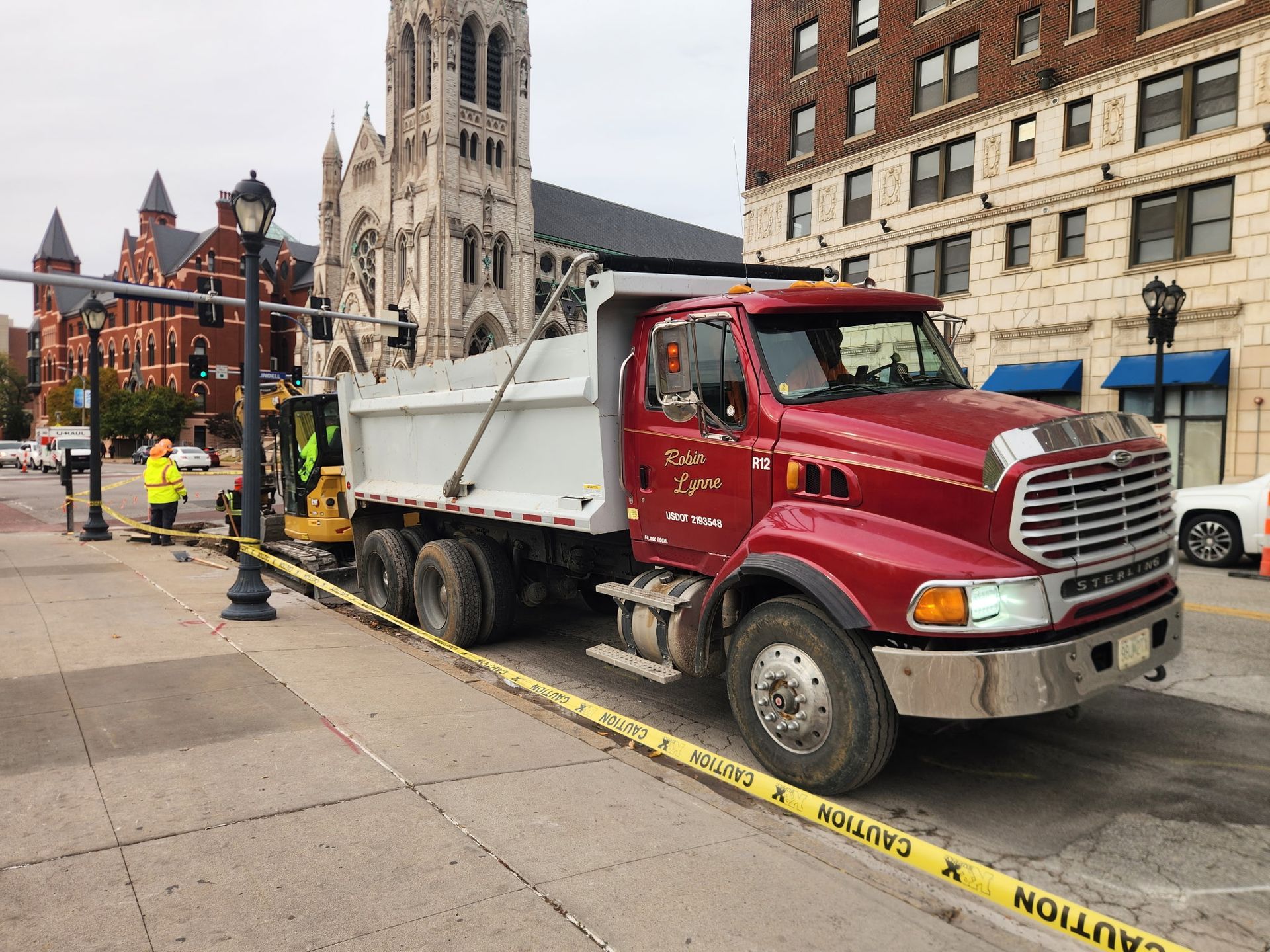 Dump truck and construction worker on a city street, caution tape, church and brick buildings in background.