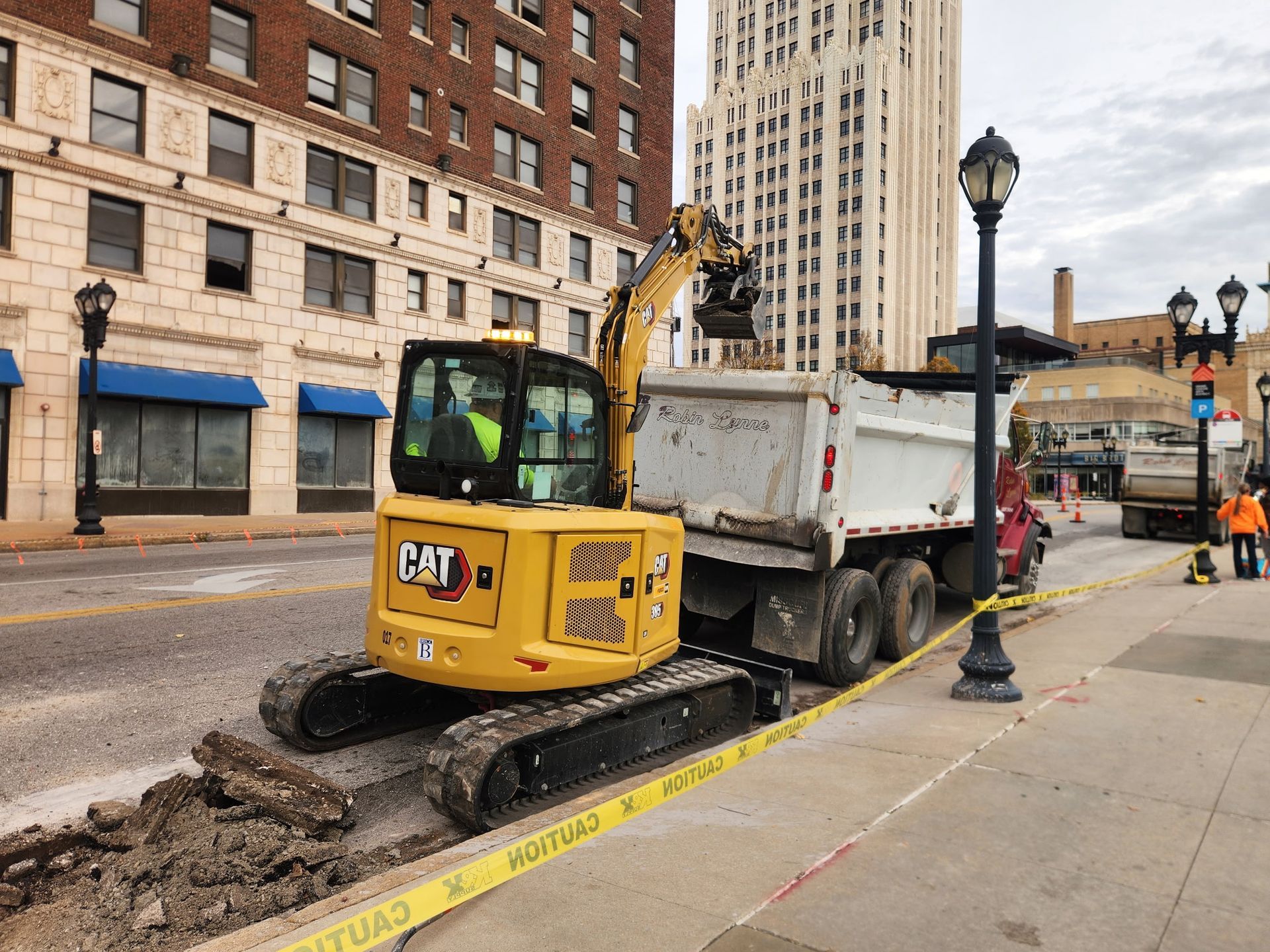 Yellow CAT excavator loading dirt into a dump truck on a city street. Buildings in background.