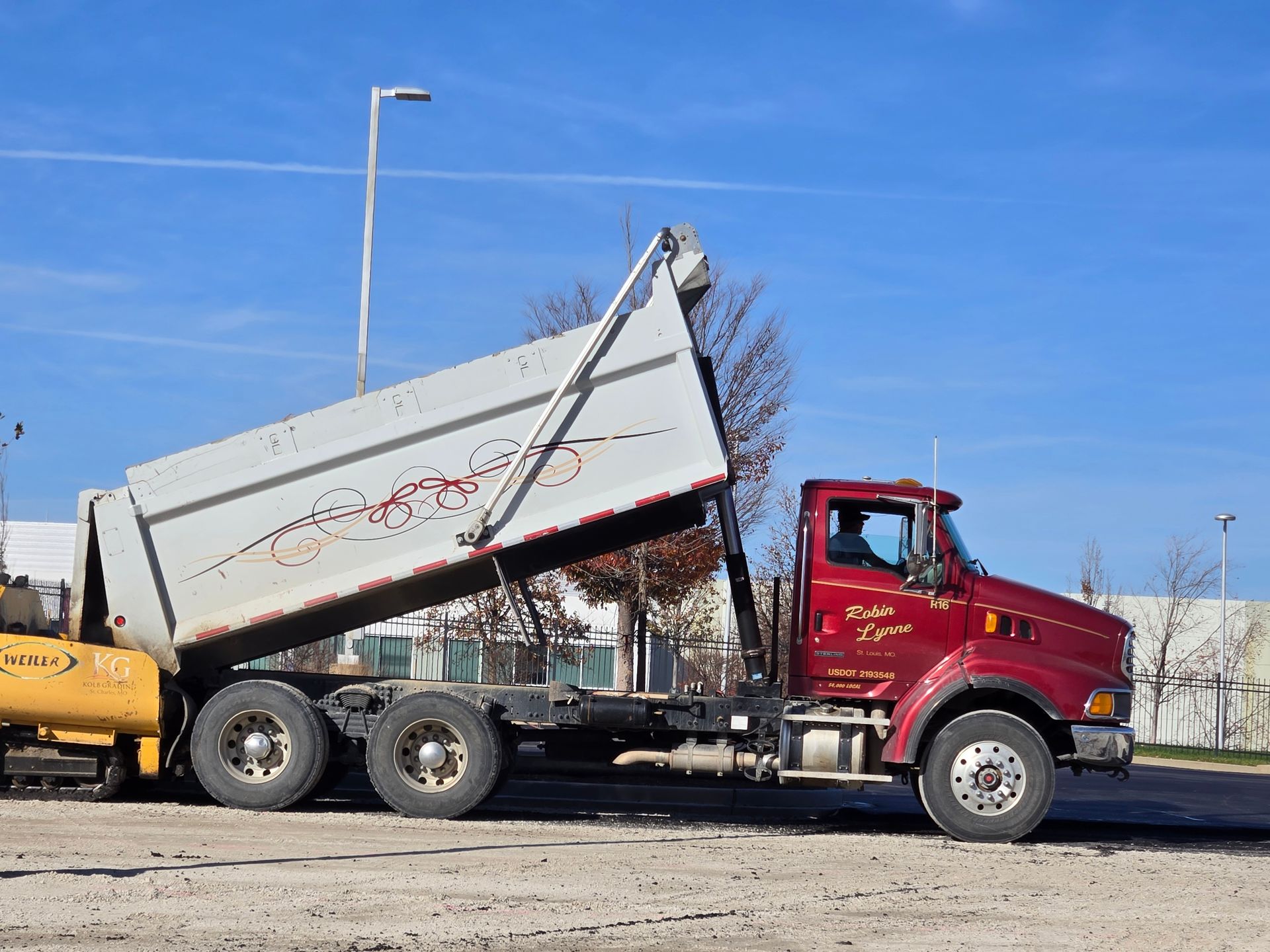 Dump trucks parked in a snowy lot at sunset. Pink and orange sky with bare trees.