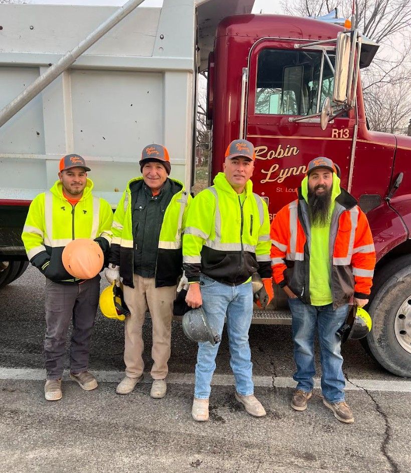 Four construction workers in safety vests stand beside a red dump truck, likely after a job.