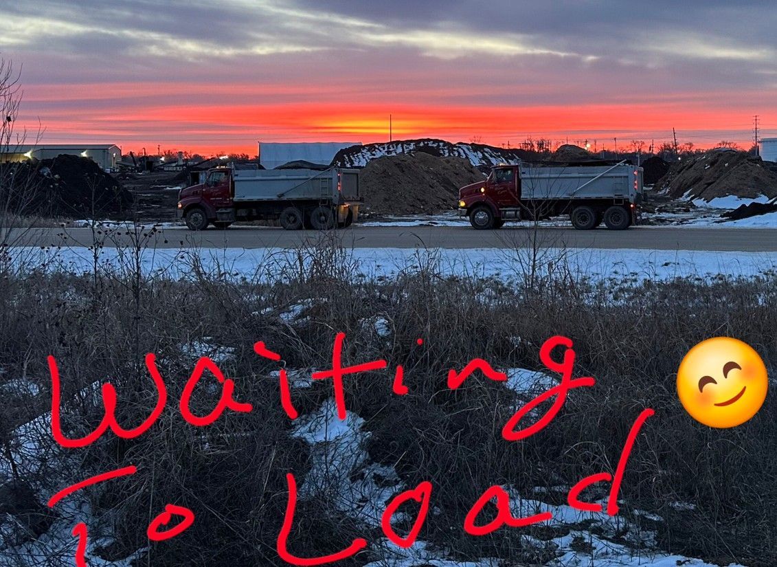 Two dump trucks wait to load at sunrise; red and orange sky, snow on the ground.