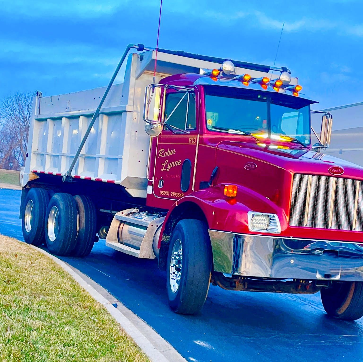 Red dump truck on a road, gray bed raised, clear sky in the background.