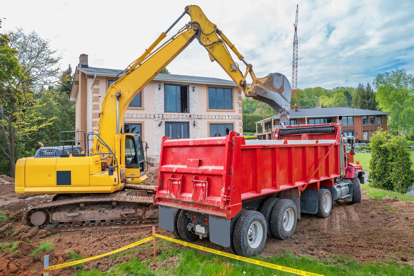 Yellow excavator loading red dump truck at a house construction site.