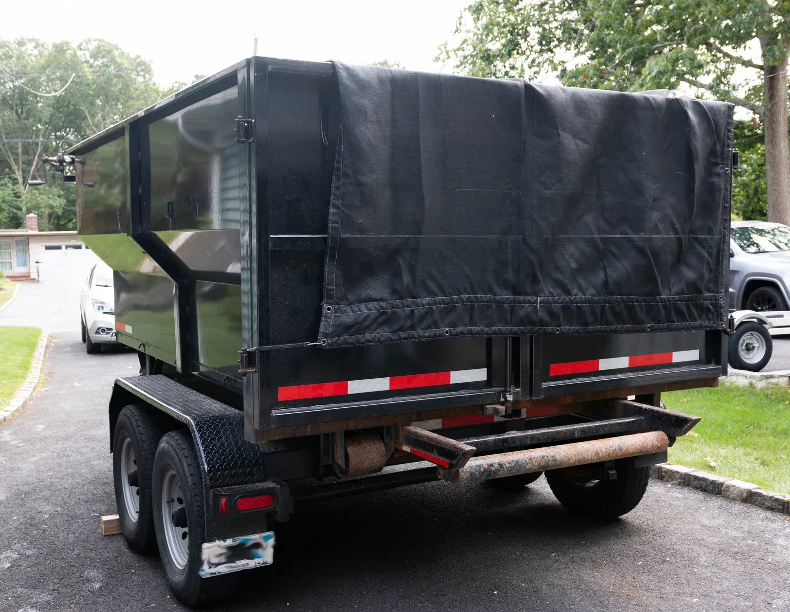 Rear view of a black trash trailer with a tarp cover. Reflective red and white stripes on bumper.