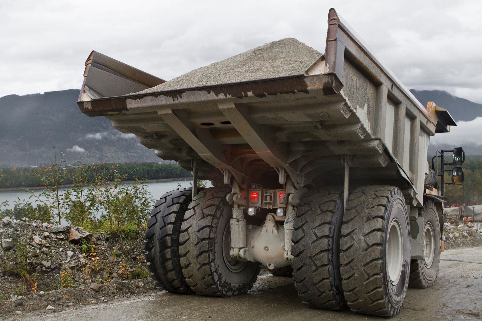 Dump truck loaded with gravel, parked on a dirt road with mountains and water in the background.