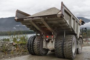 Dump truck loaded with gravel, parked on a dirt road with mountains and water in the background.