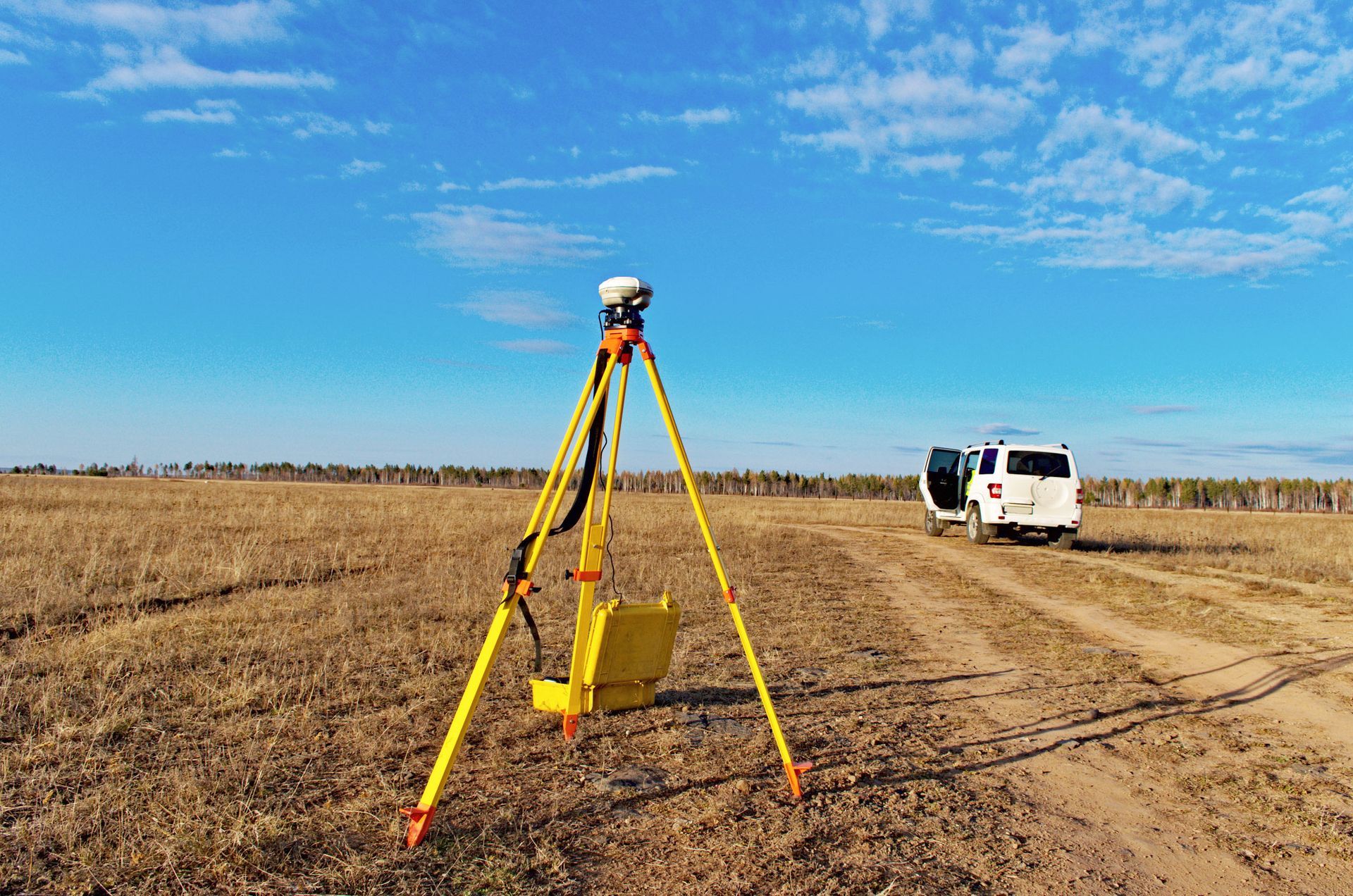 A GPS surveying setup on a tripod in a field with a white vehicle parked nearby, under a blue sky.