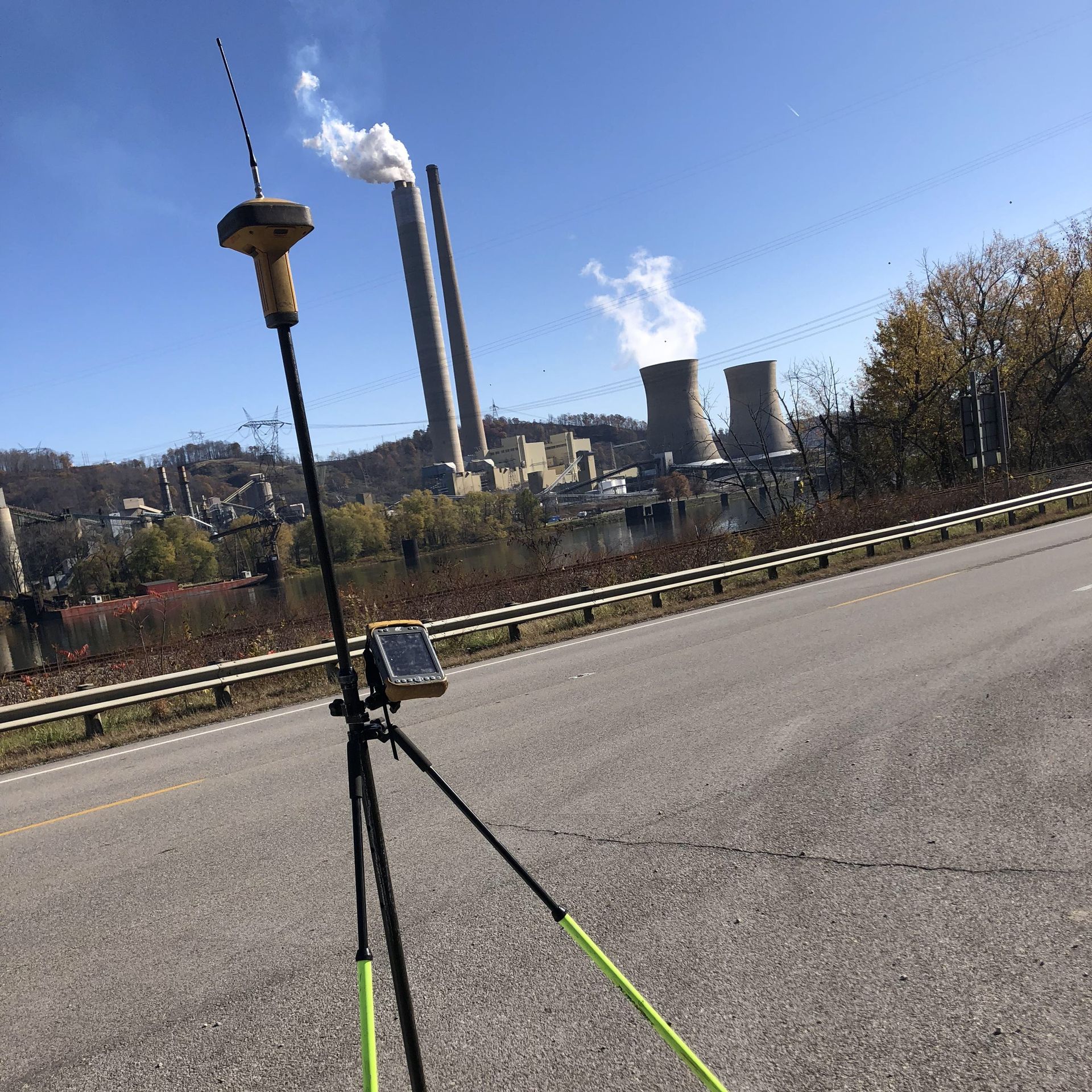 GPS surveying equipment set up on a road, with a power plant in the background on a sunny day.