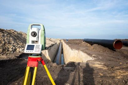 A surveying instrument on a tripod in a trench, likely measuring for pipeline construction. Dark pipes and dirt are visible.