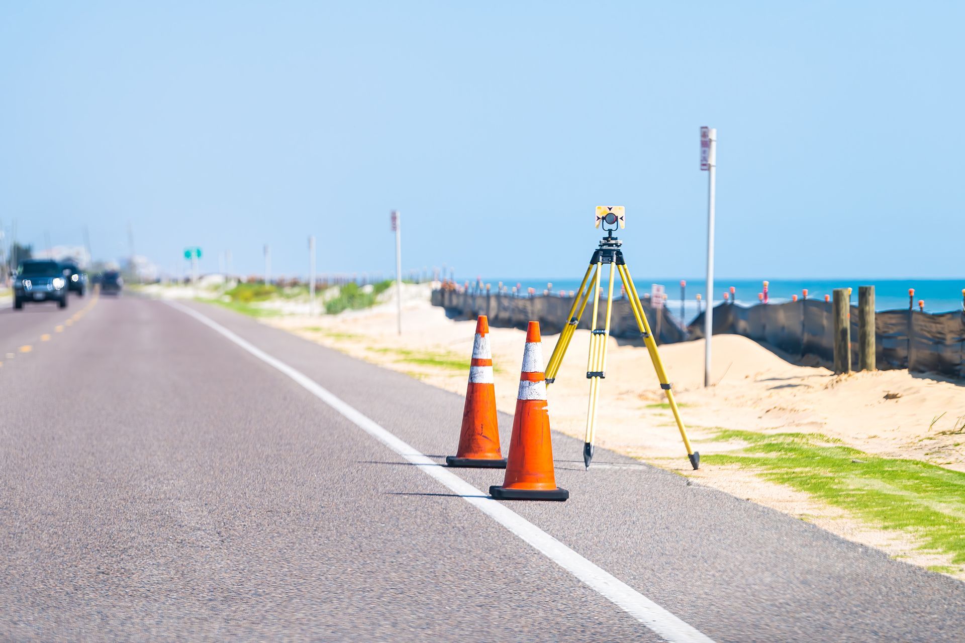 A road with orange traffic cones and surveying equipment, next to a beach with the ocean in the background.
