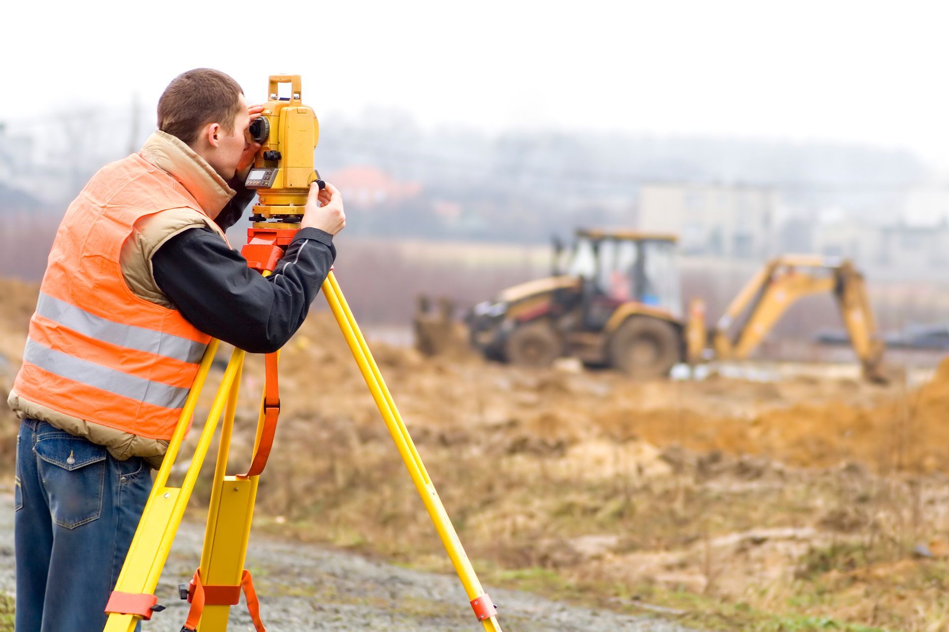 A surveyor in an orange vest uses a theodolite on a tripod at a construction site. A bulldozer is blurred in the background.