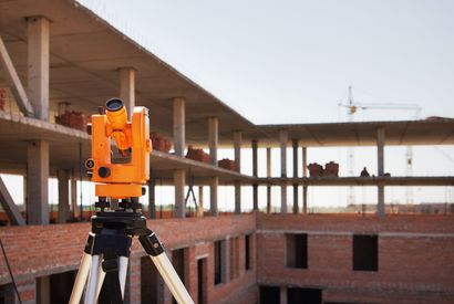 A surveying instrument on a tripod at a construction site; orange instrument, brick building in progress, sunny day.